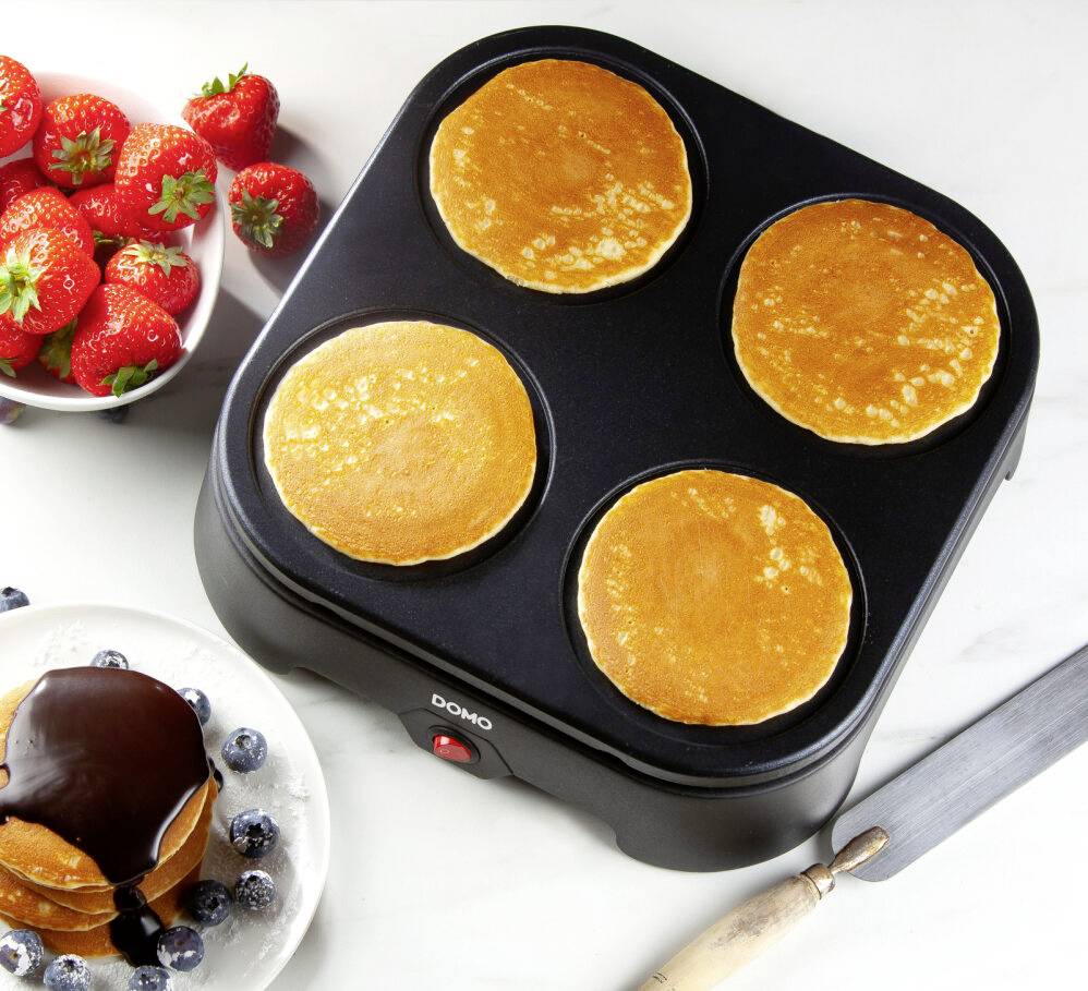 Four fresh pancakes on an electric pancake maker. Beside it a bowl of strawberries and a plate with pancakes and blueberries.