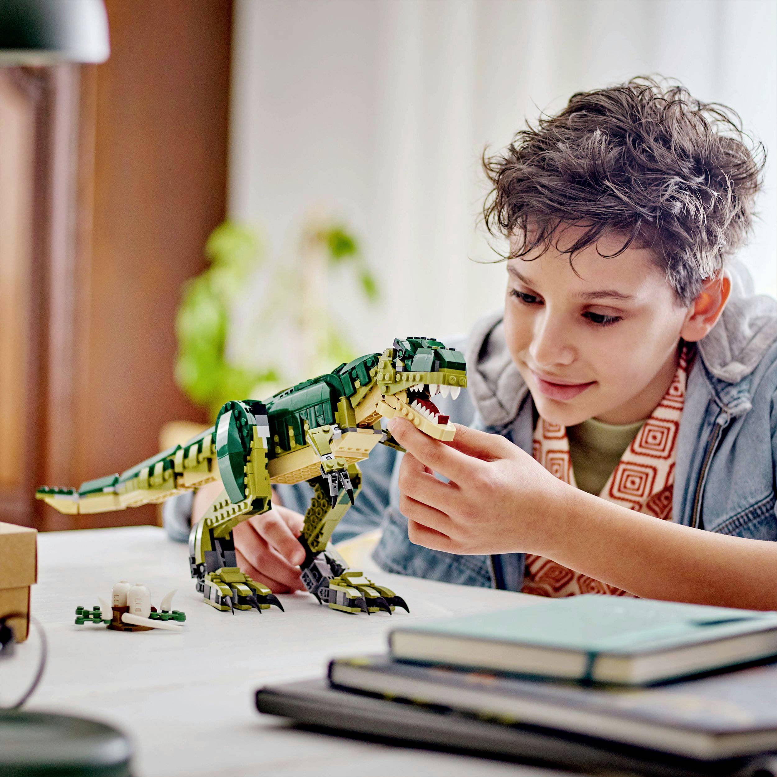 A child is playing joyfully with a Lego dinosaur on a table, next to a notebook and some plants.
