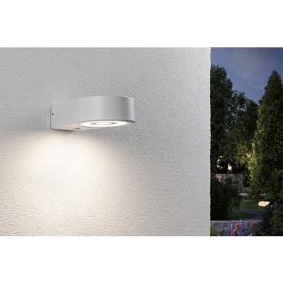 Modern, round outdoor light mounted on a white wall, casting a warm glow. Trees and a garden are visible in the background at dusk.