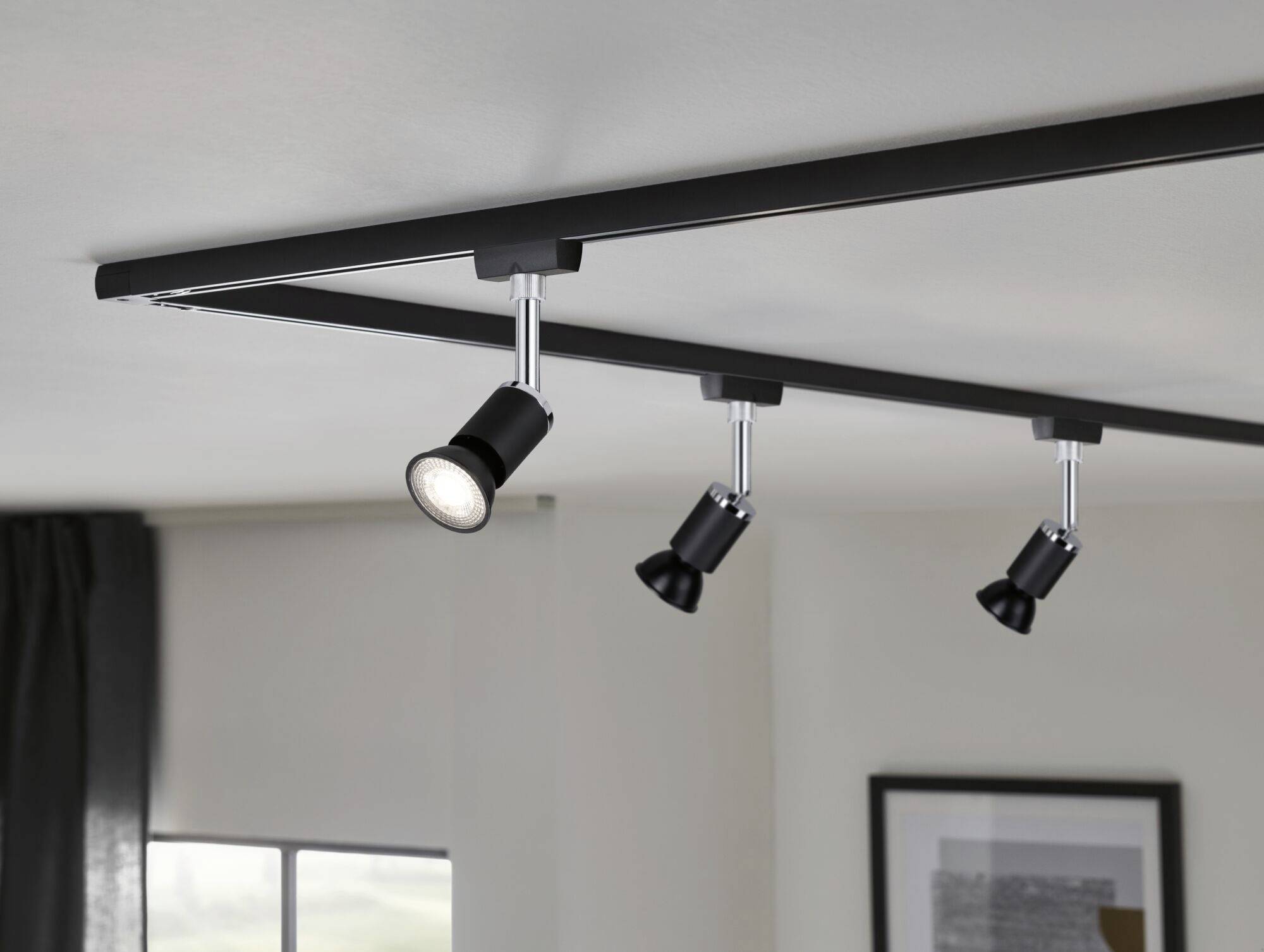 Black ceiling track lighting with four adjustable spotlights, mounted on a white ceiling in a modern room.