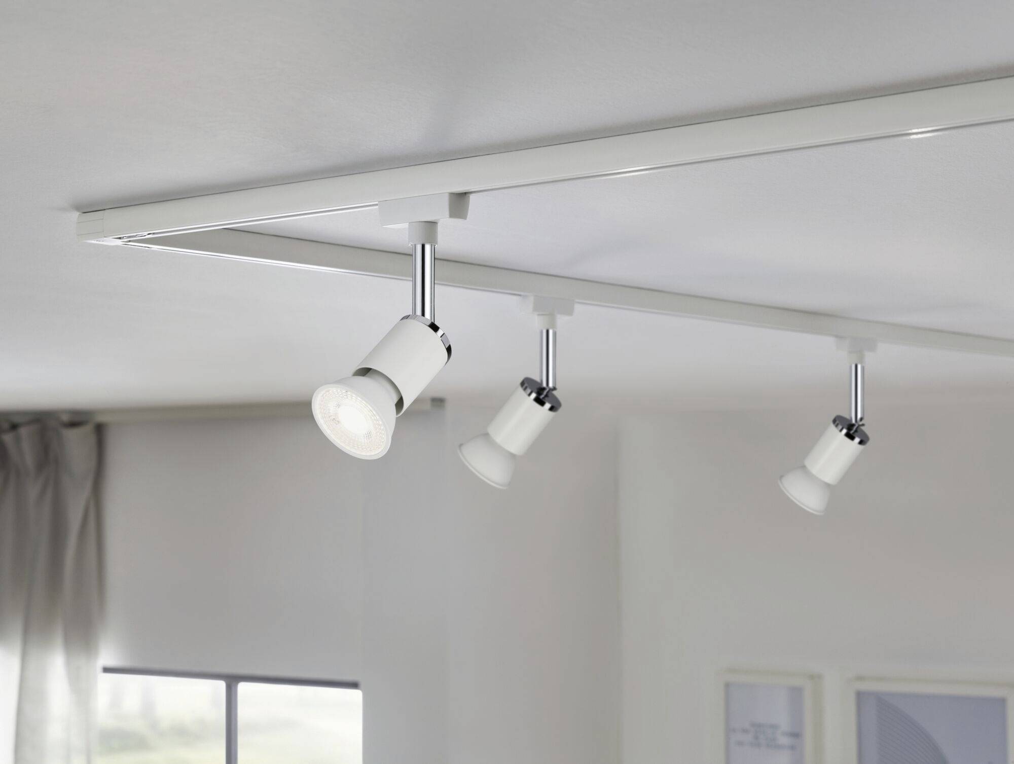 Three white ceiling spotlights on a track mounted to the ceiling, pointing downwards. Window and curtain in the background.