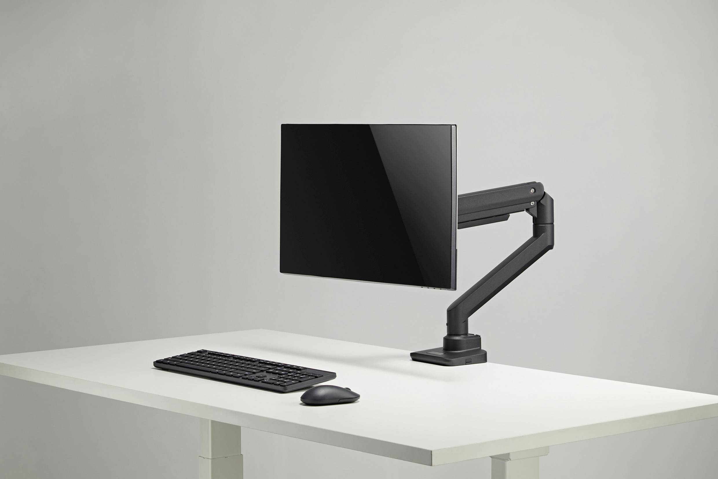 A computer monitor on an adjustable arm is attached to a white desk. Next to it are a keyboard and a mouse.