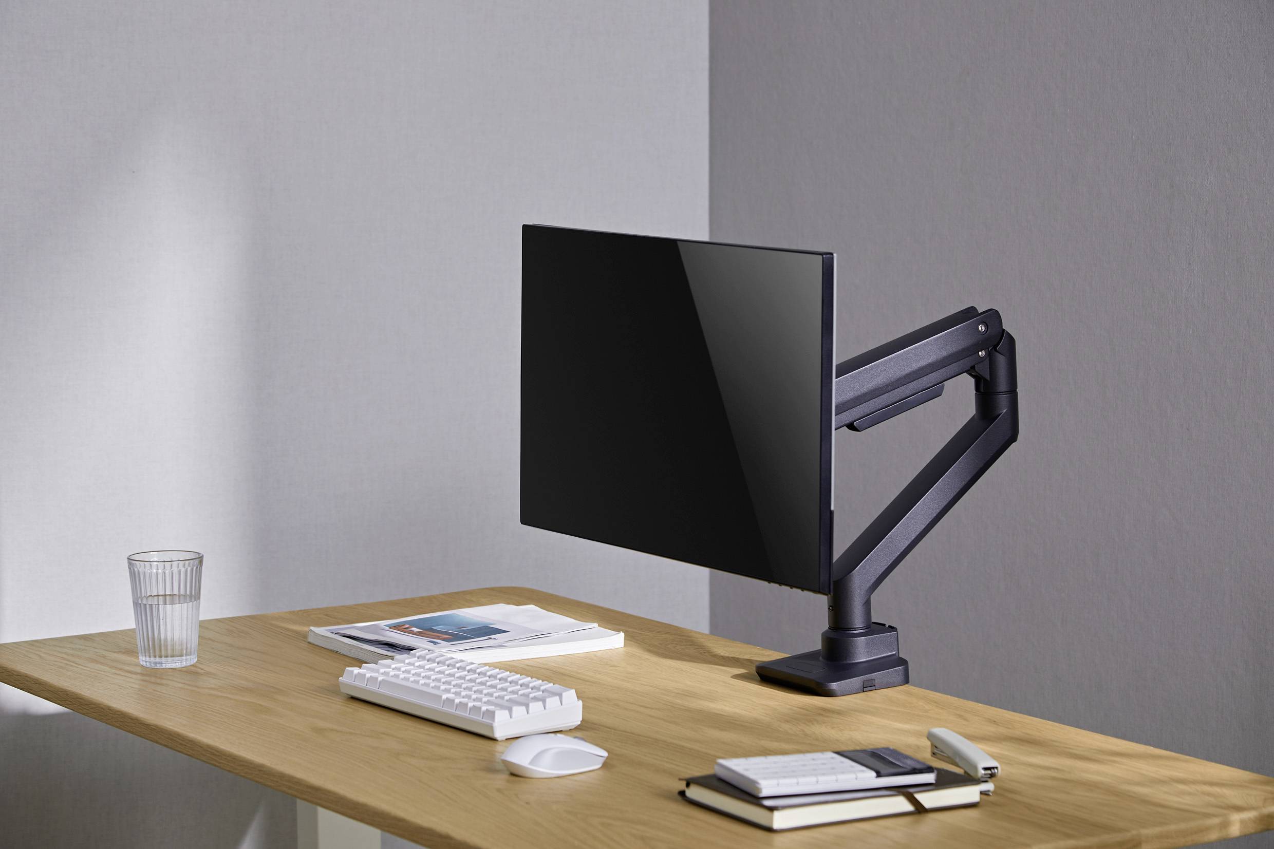 A desk with a monitor on an adjustable arm, keyboard, mouse, notebooks, and a glass. Modern, minimalist setup.