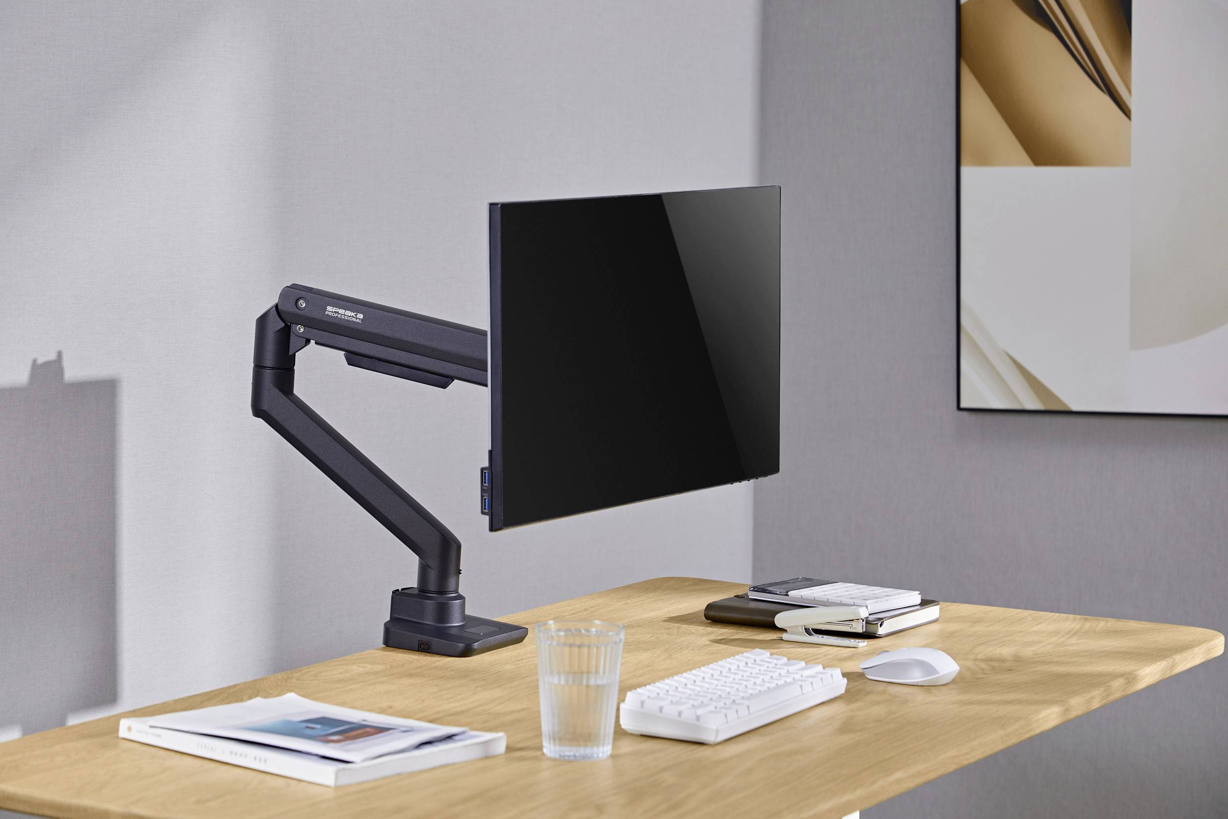 A wooden desk with a black monitor on an adjustable arm. Sitting on it are a keyboard, mouse, glass of water and two books.
