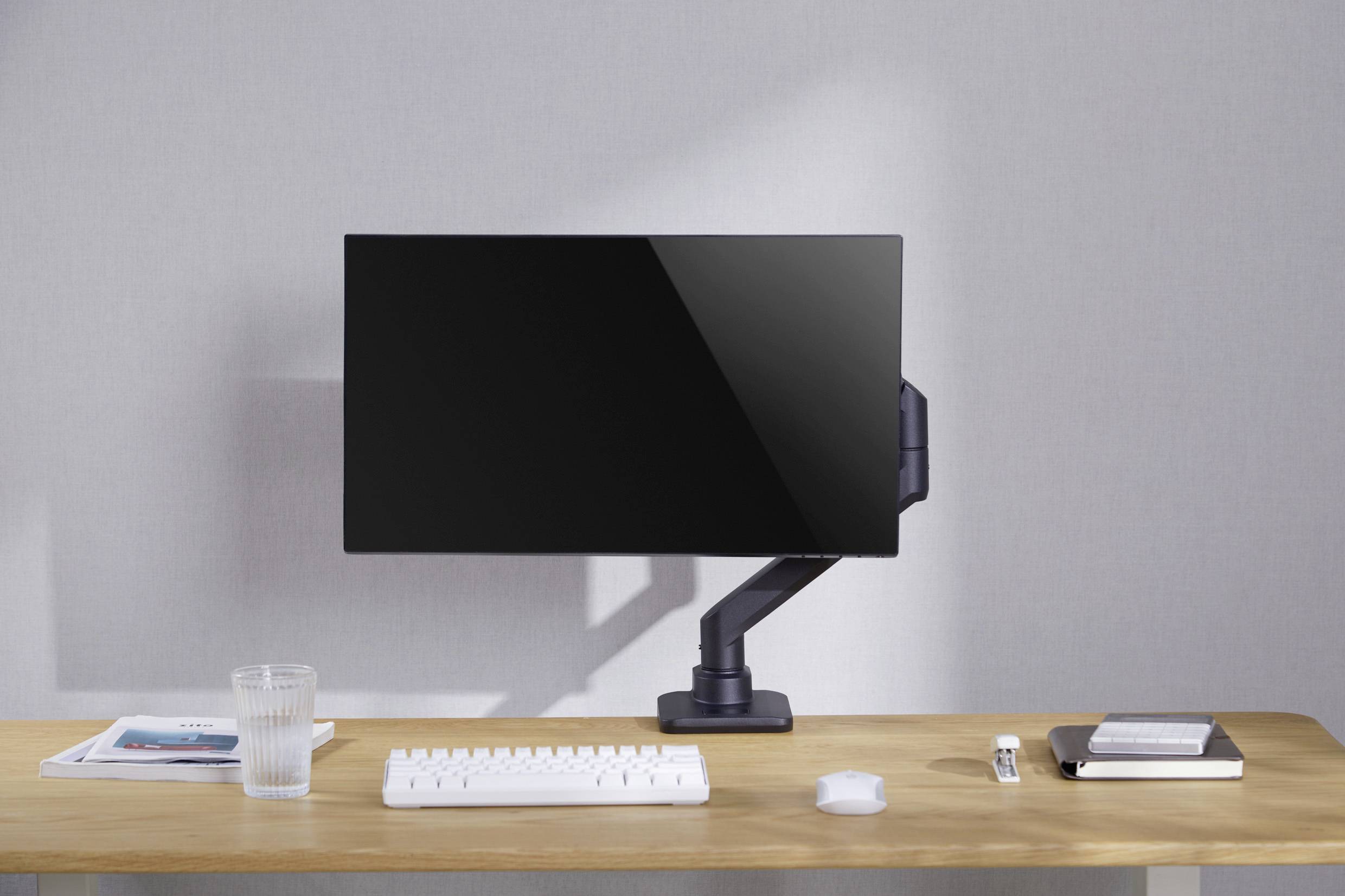 A desk with a monitor on a stand, beside it a keyboard, mouse, glass, remote control, notebook and magazine, in front of a grey wall.
