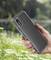 A hand is holding a smartphone with a transparent case in front of a blurry, green woodland landscape with a lake in the background.