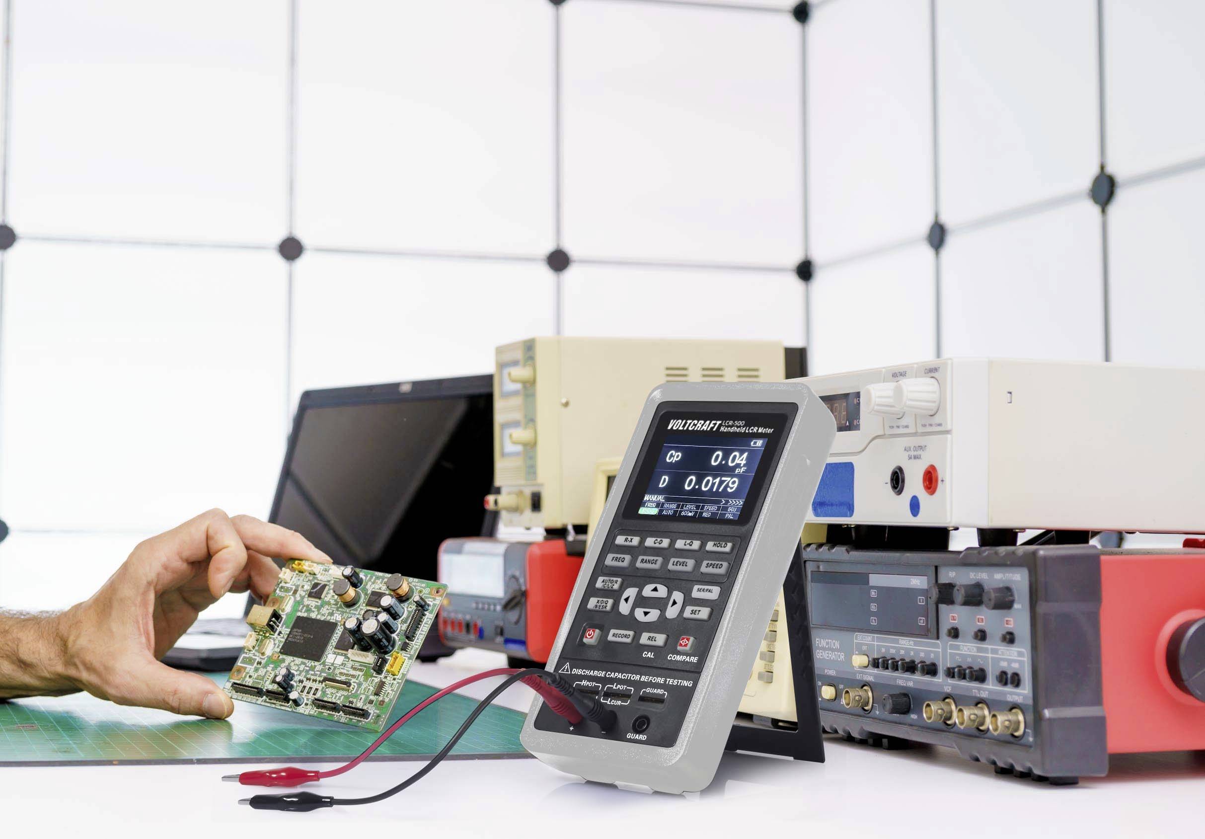 A person is holding a circuit board next to several electronic measuring instruments on a table. This is a laboratory setting.