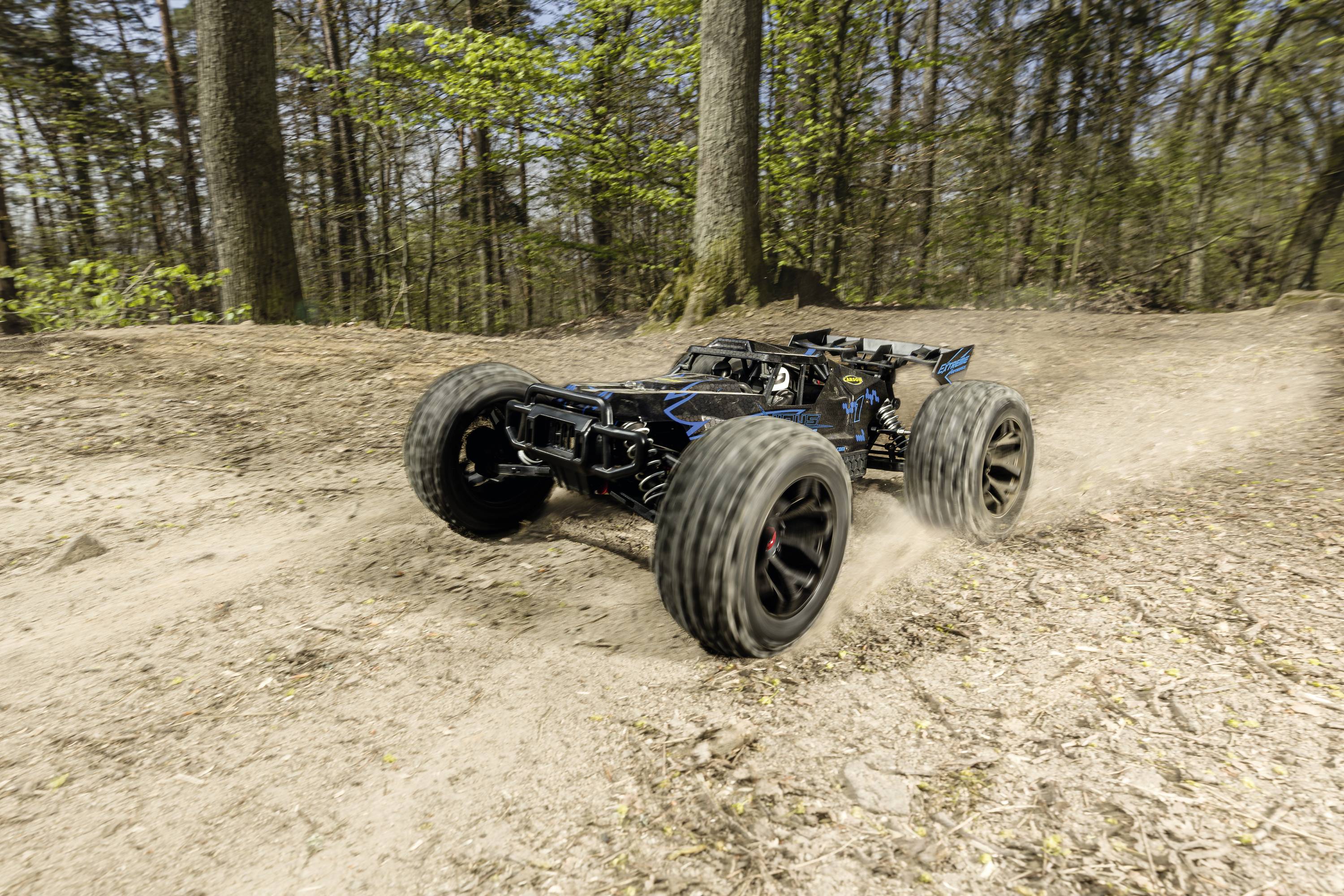 Remote-controlled car drives quickly along a sandy forest track. The large tyres kick up dust. Trees form the background.