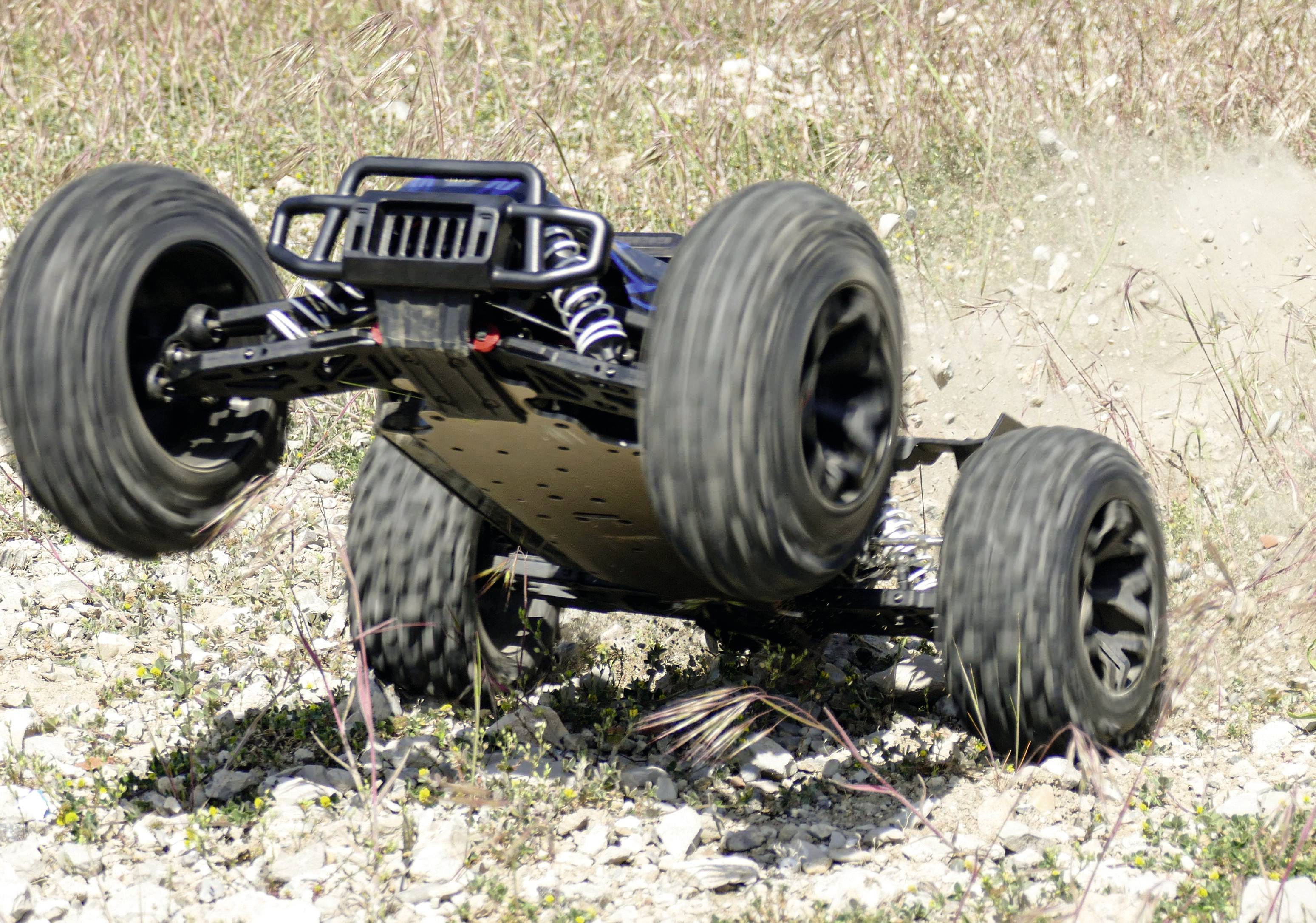 A remote-controlled car speeds across a rocky outdoor surface, lifting its front wheels off the ground.