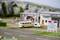 A beige model car with a trailer is parked on a diorama in front of a petrol station. In the background, models of buildings and trees can be seen.