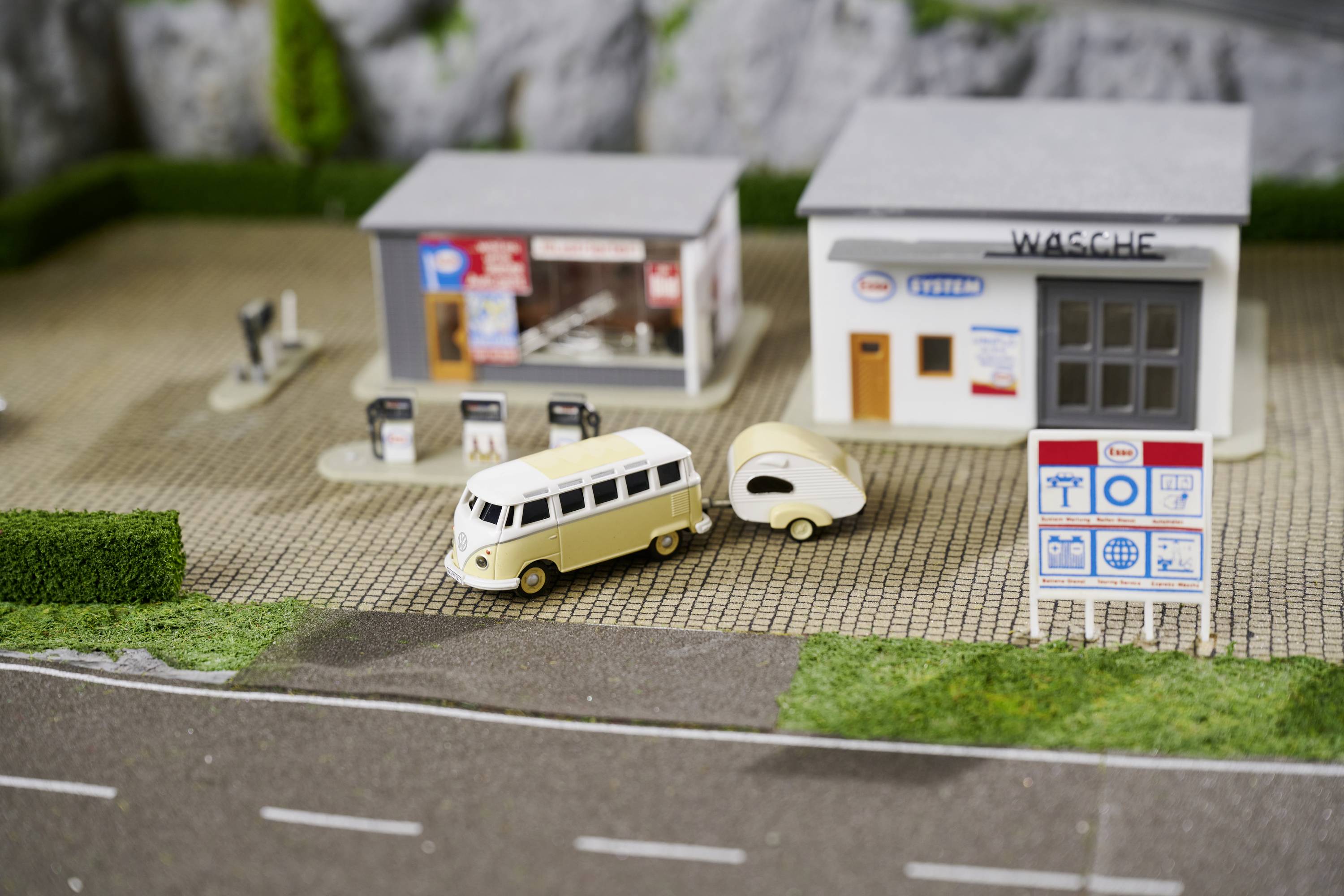 A model car with a trailer is parked in front of a miniature petrol station. Next to the petrol station are small buildings and an advertising sign.
