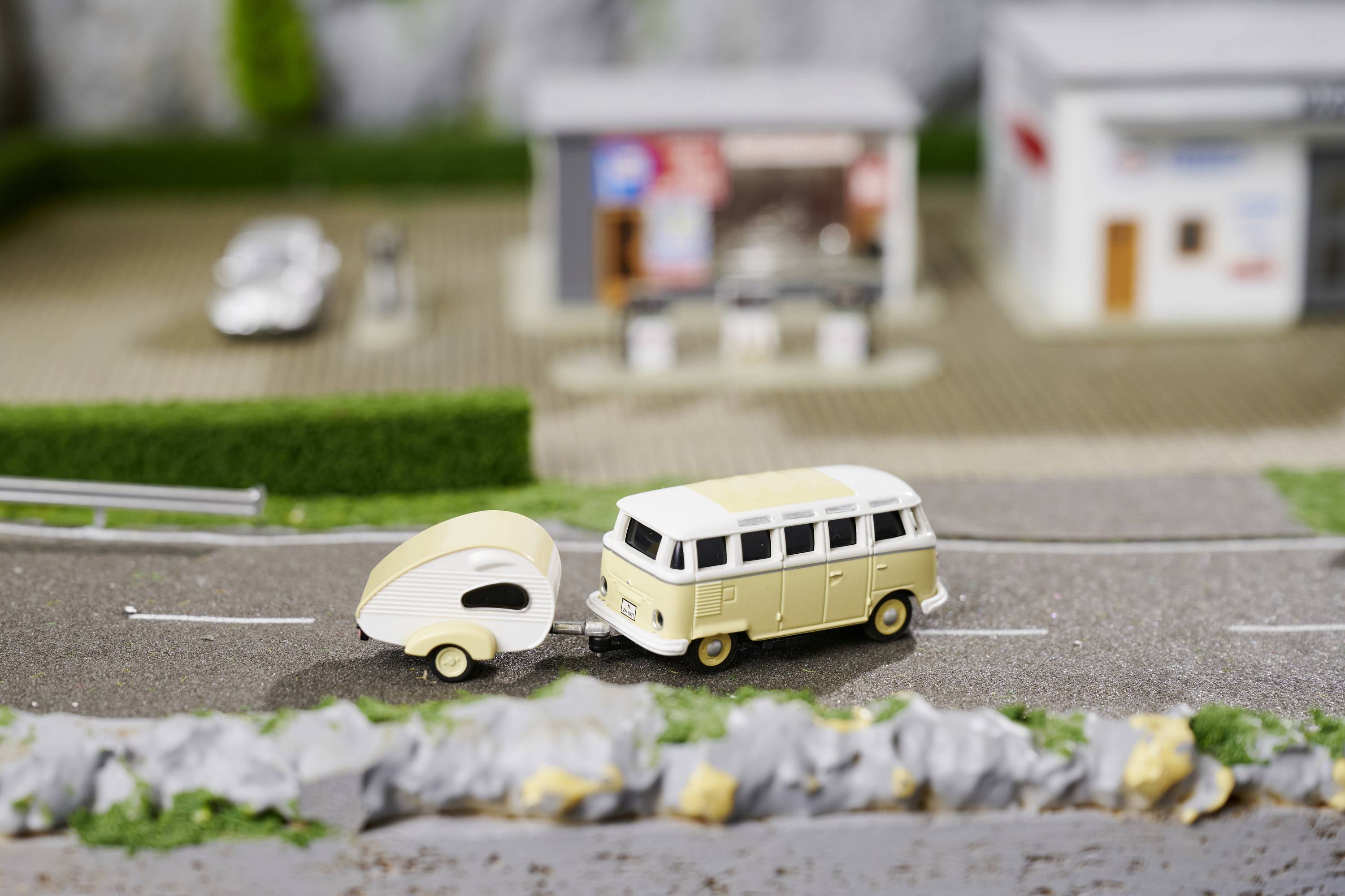 A vintage-style model car with a small trailer drives along a miniature road past a model building.