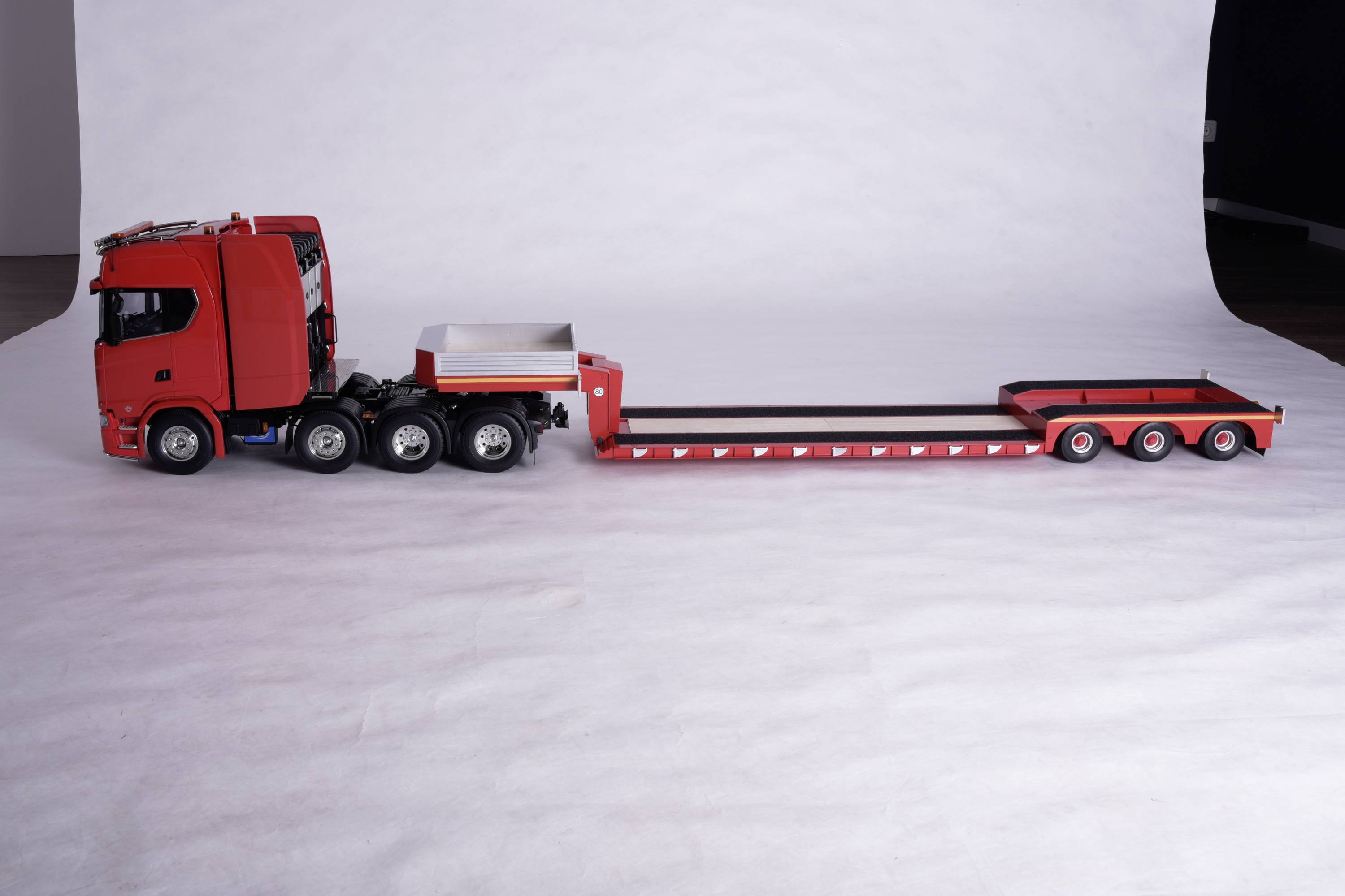 A red lorry with a long, flat trailer is parked on a white surface.