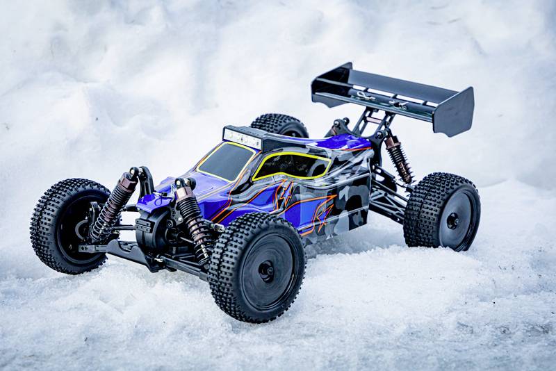 A blue and yellow remote-controlled car with black wheels is standing in the snow. It has a large rear spoiler for stability.