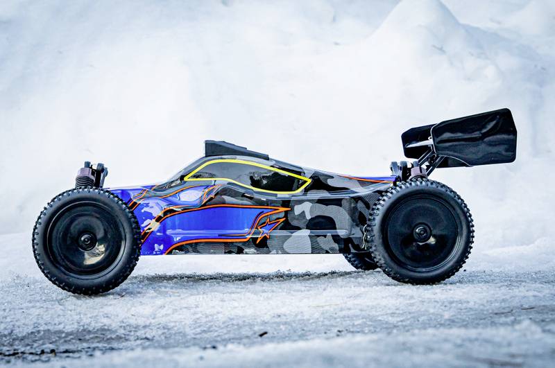 A remote-controlled car with a blue and black, camouflage-patterned chassis, stands on snow-covered ground. It has large tyres.