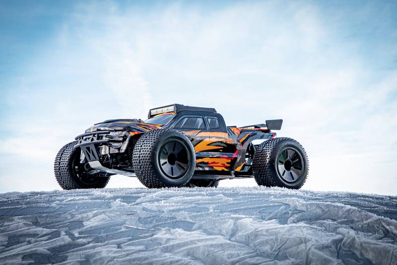 A remote-controlled car with an orange and black design stands on an icy, snow-covered surface beneath a blue sky.