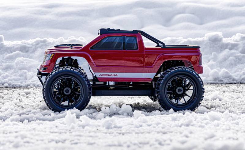 A red remote-controlled car sits on snow-covered ground, surrounded by snowdrifts in the background.