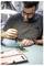 A person with glasses is focused on using a screwdriver to work on a circuit board at a desk, under a lit lamp. A tool case is open nearby.