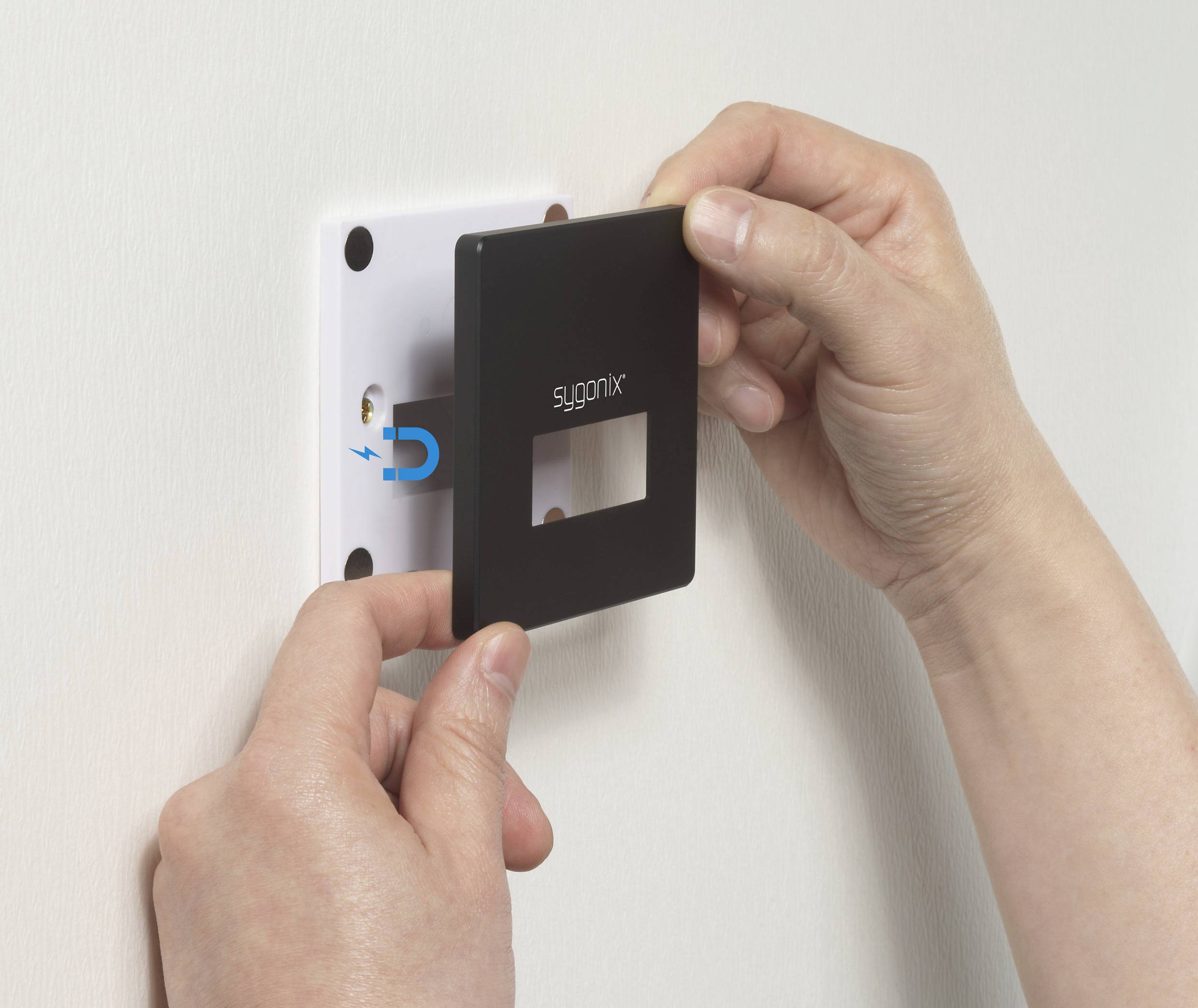 'A person is attaching a black, square front plate with the inscription "sygonix" to a socket holder on the wall.'