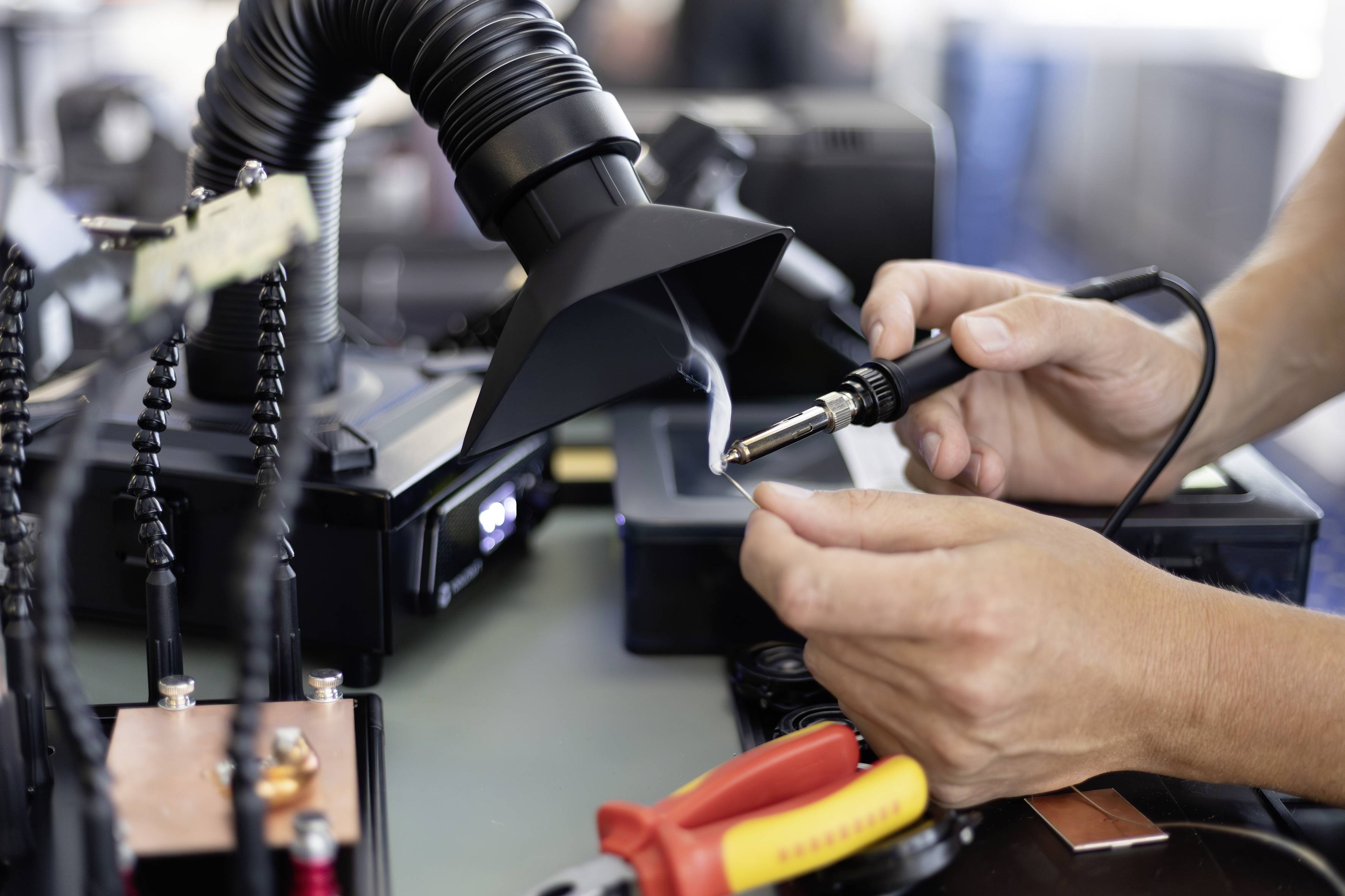 Hands using a soldering iron to repair a circuit board under a fume extractor, with electronics tools on the workbench.