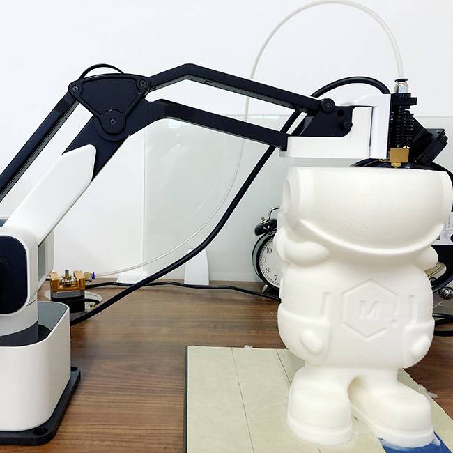 A robotic arm is printing a white 3D figure on a table. In the background, an alarm clock and another mechanical device can be seen.