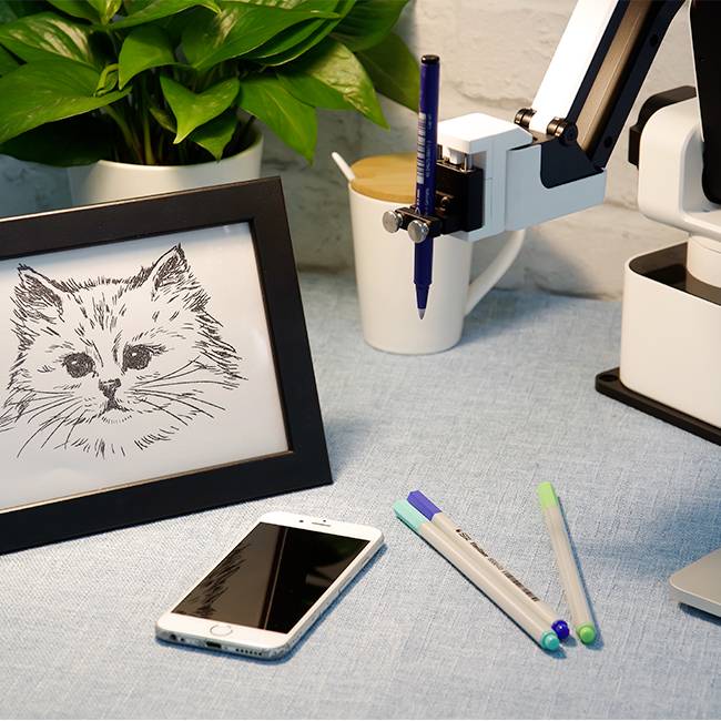 A robotic arm is drawing a cat picture on a table. Next to it are pens, a mobile phone and a potted plant.