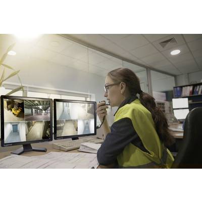 A woman wearing a yellow high-visibility vest is sitting at a desk and monitoring video camera feeds on two screens in a control room.