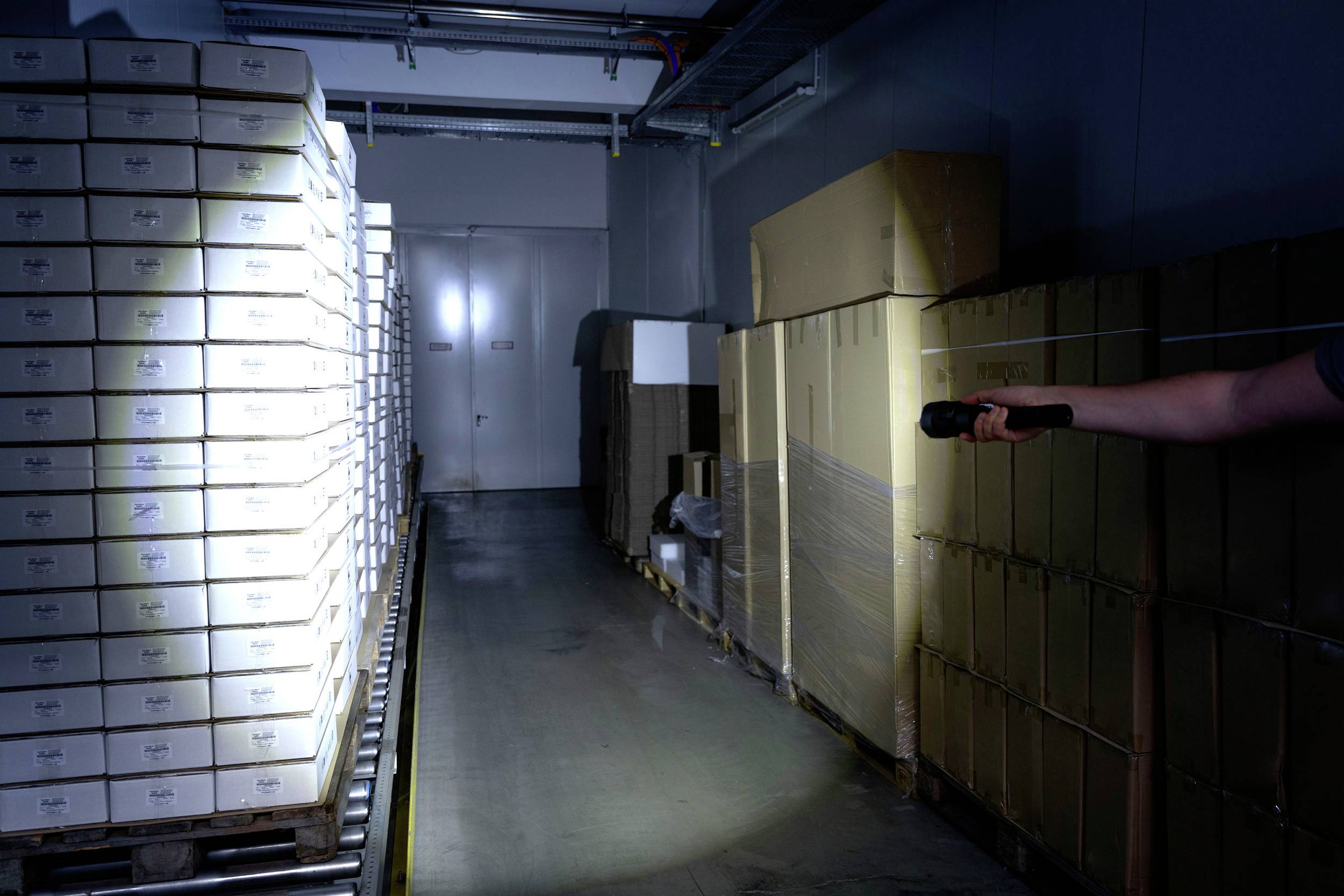 In a dimly lit storage room, a person with a torch points towards stacked boxes that form a narrow passageway.