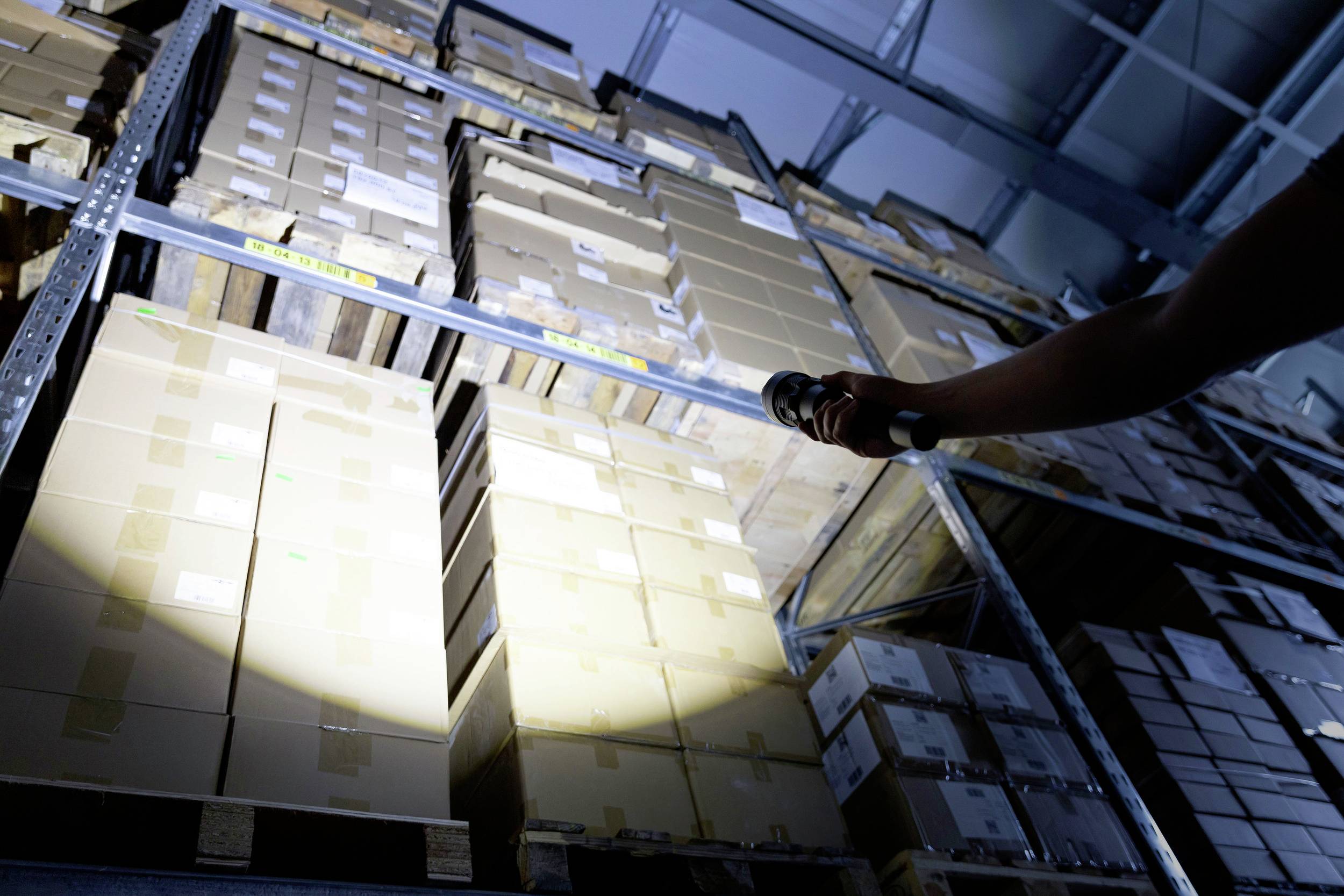 A person is illuminating a row of stacked boxes in a large storage room with a torch.