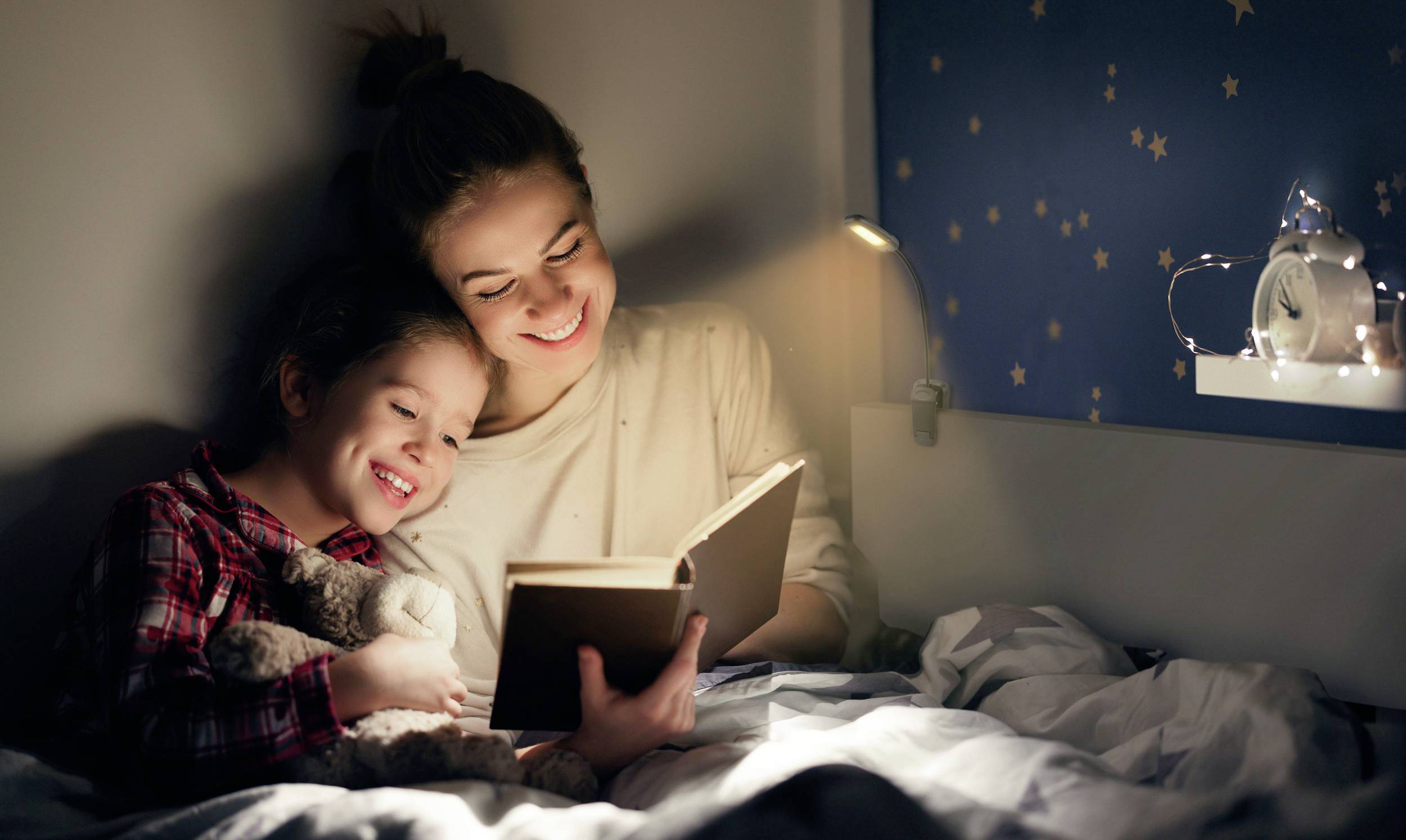 A woman and child are sitting in bed reading a book in warm light. The child is cuddling a soft toy. Stars are visible in the background.