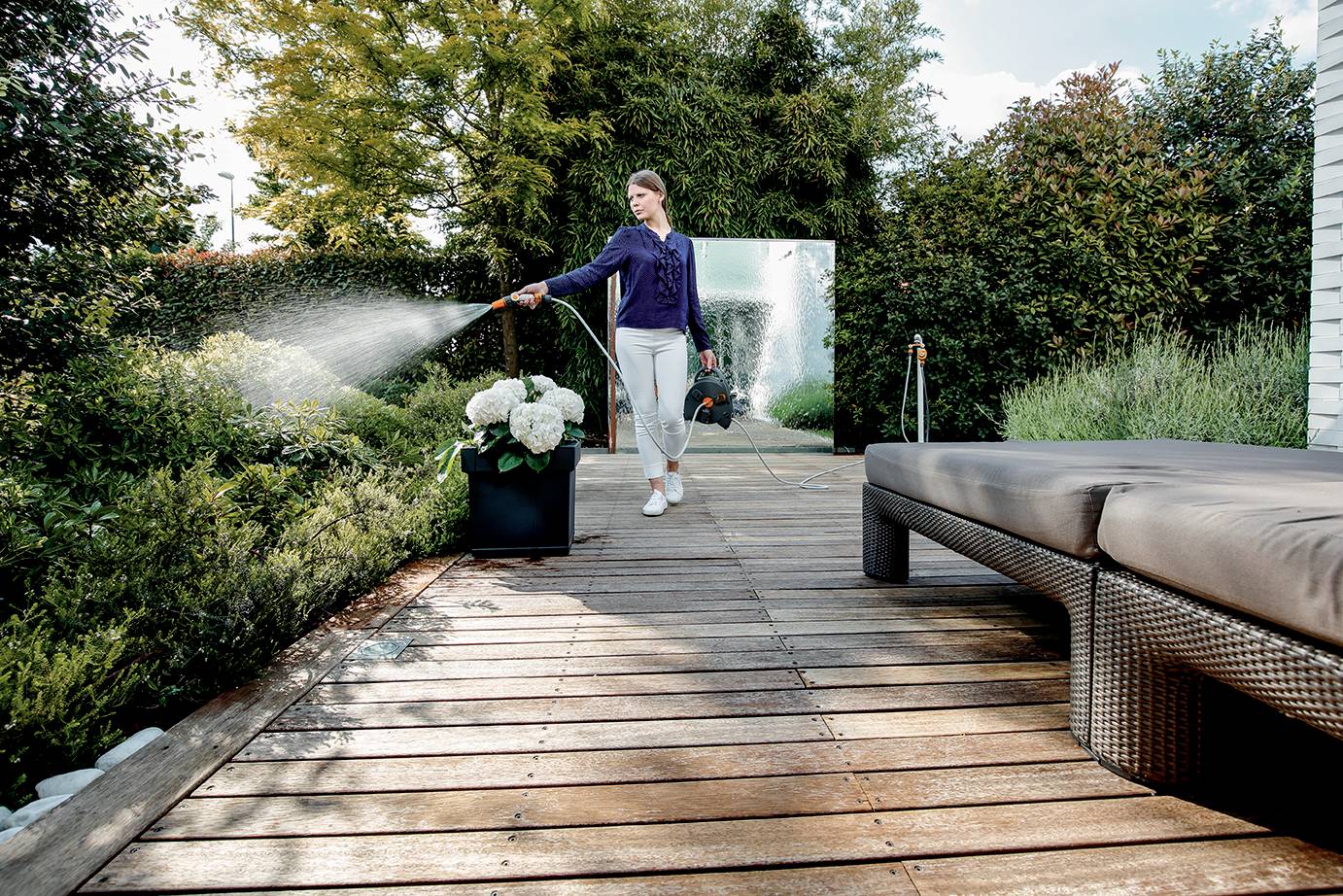 A person is watering plants with a hosepipe on a wooden terrace, surrounded by lush greenery. Bushes can be seen in the background.
