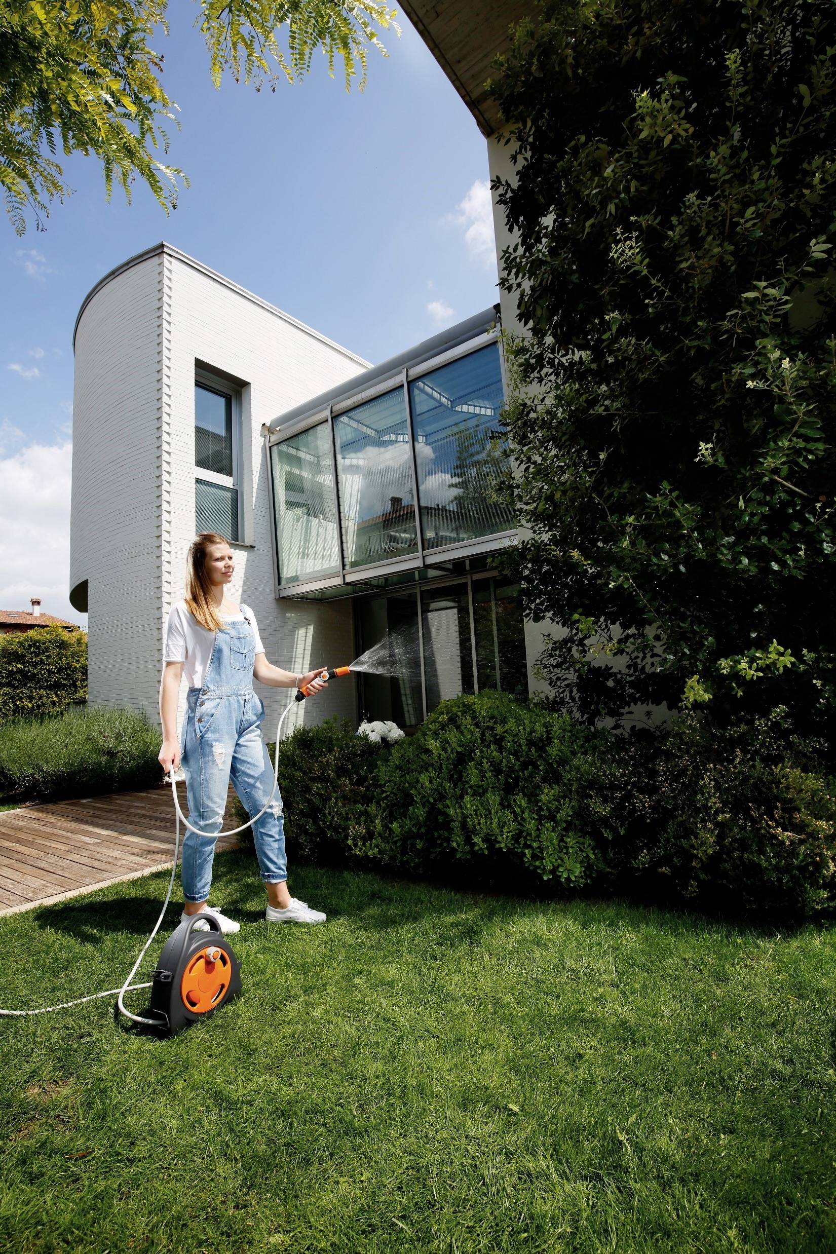 A woman is watering the lawn in front of a modern white house with large windows. She is holding a garden hose and standing on green grass.