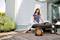 A woman sits on a terrace, surrounded by plants, reading a book. Next to her is an orange hose reel on wooden planks.