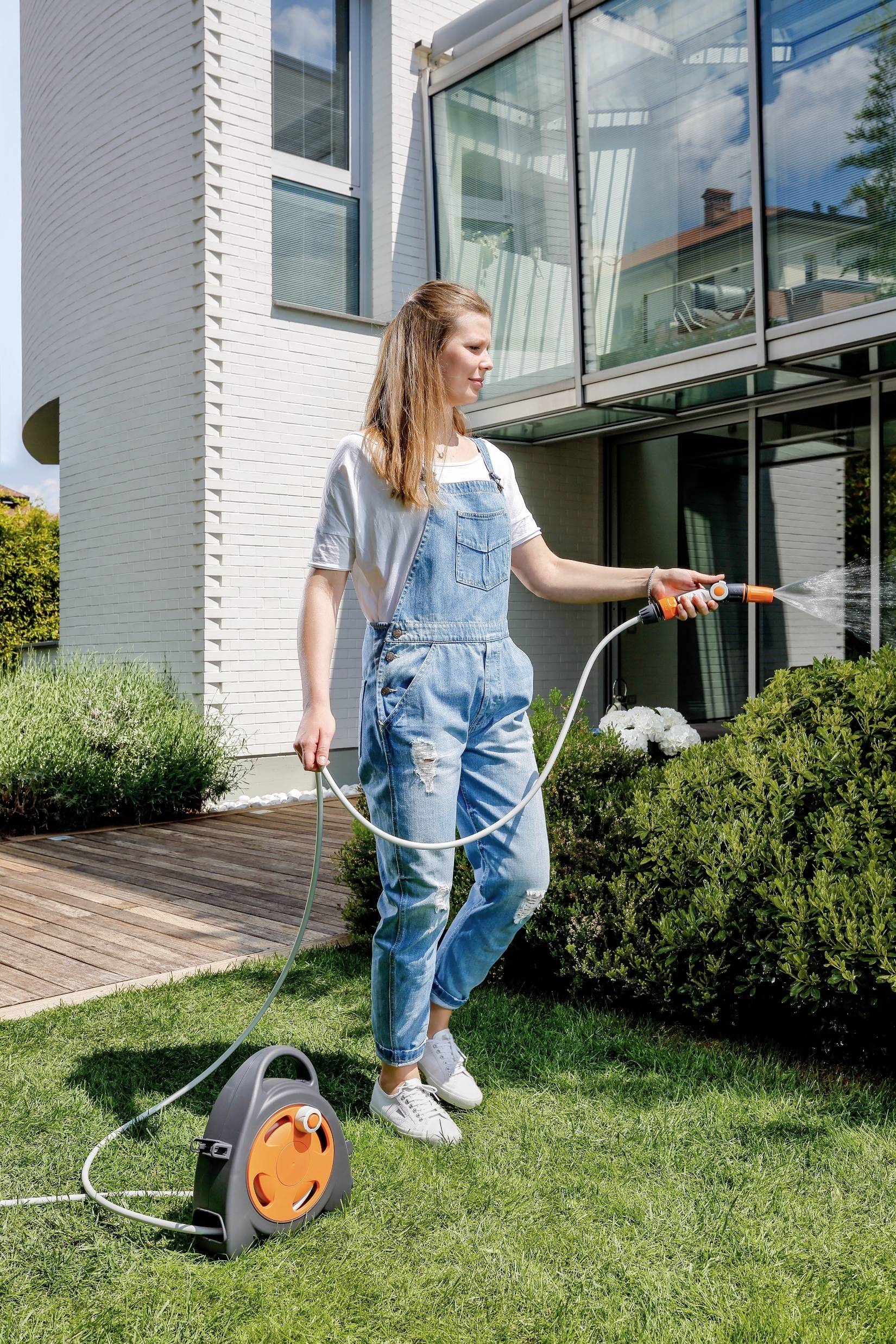 A person is watering a green lawn in front of a modern house using a hosepipe. Large windows are visible in the background.