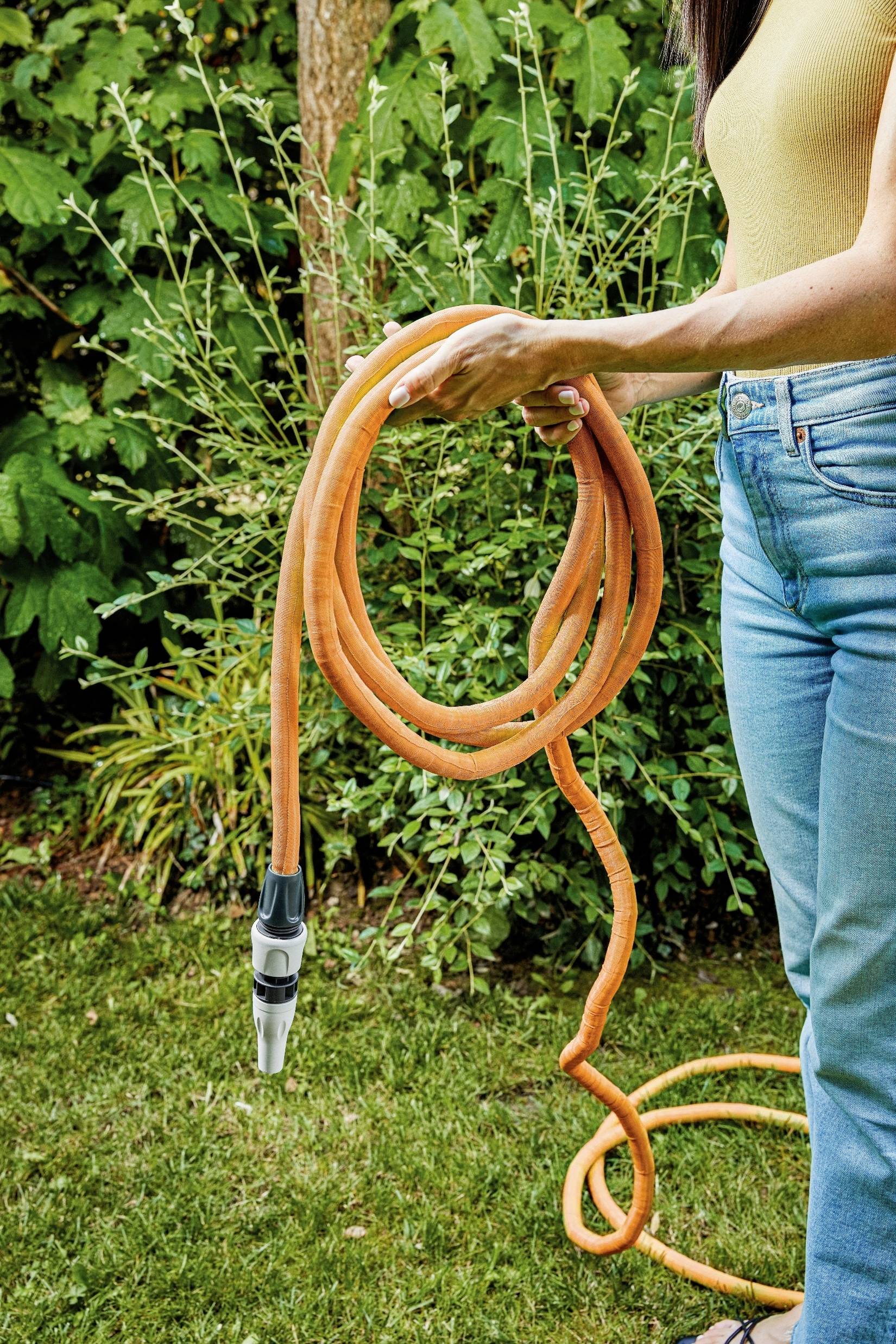 A person is holding an orange garden hose in the garden. Green bushes can be seen in the background.