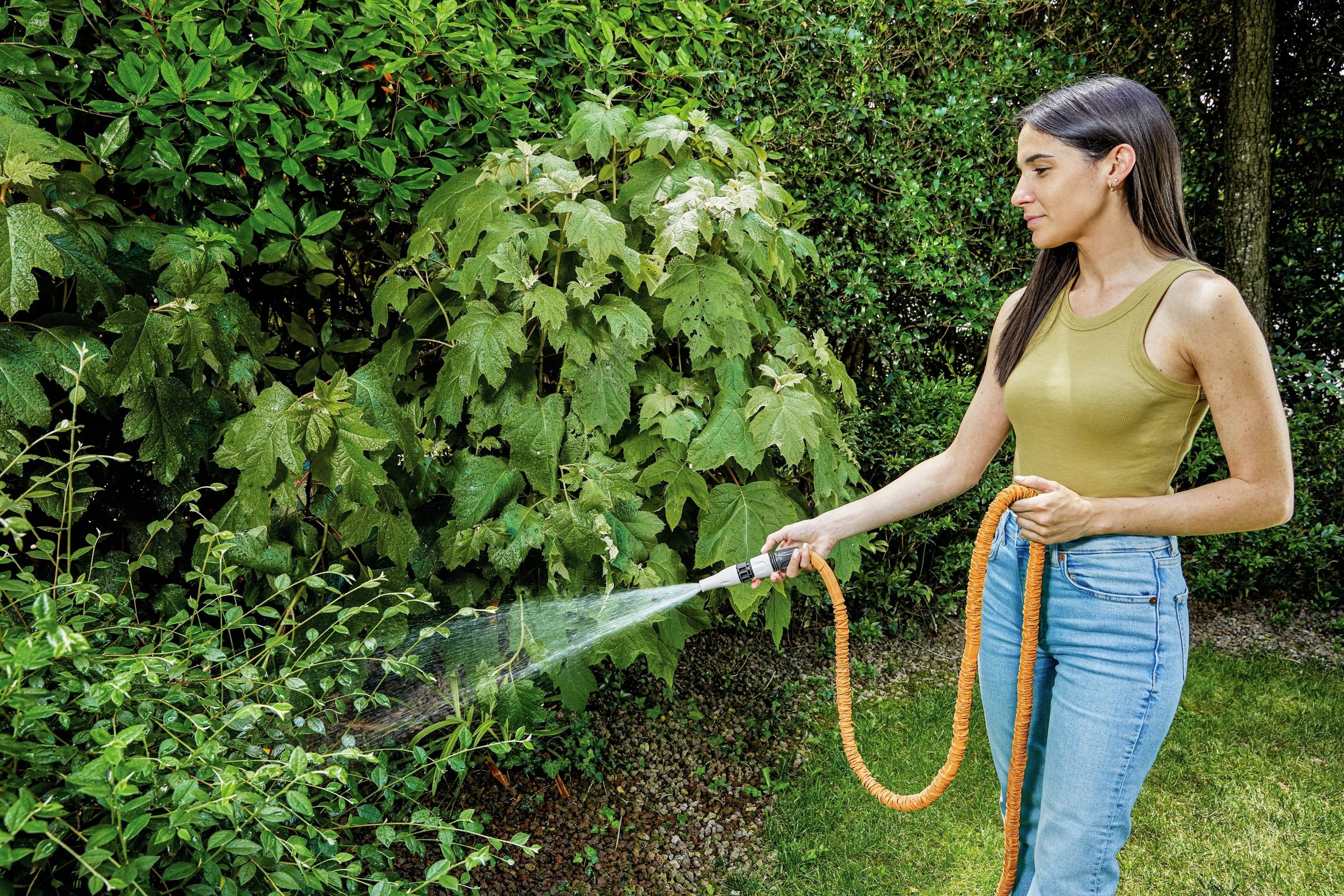 A woman is watering plants in the garden with an orange garden hose. She is wearing a beige top and blue jeans.