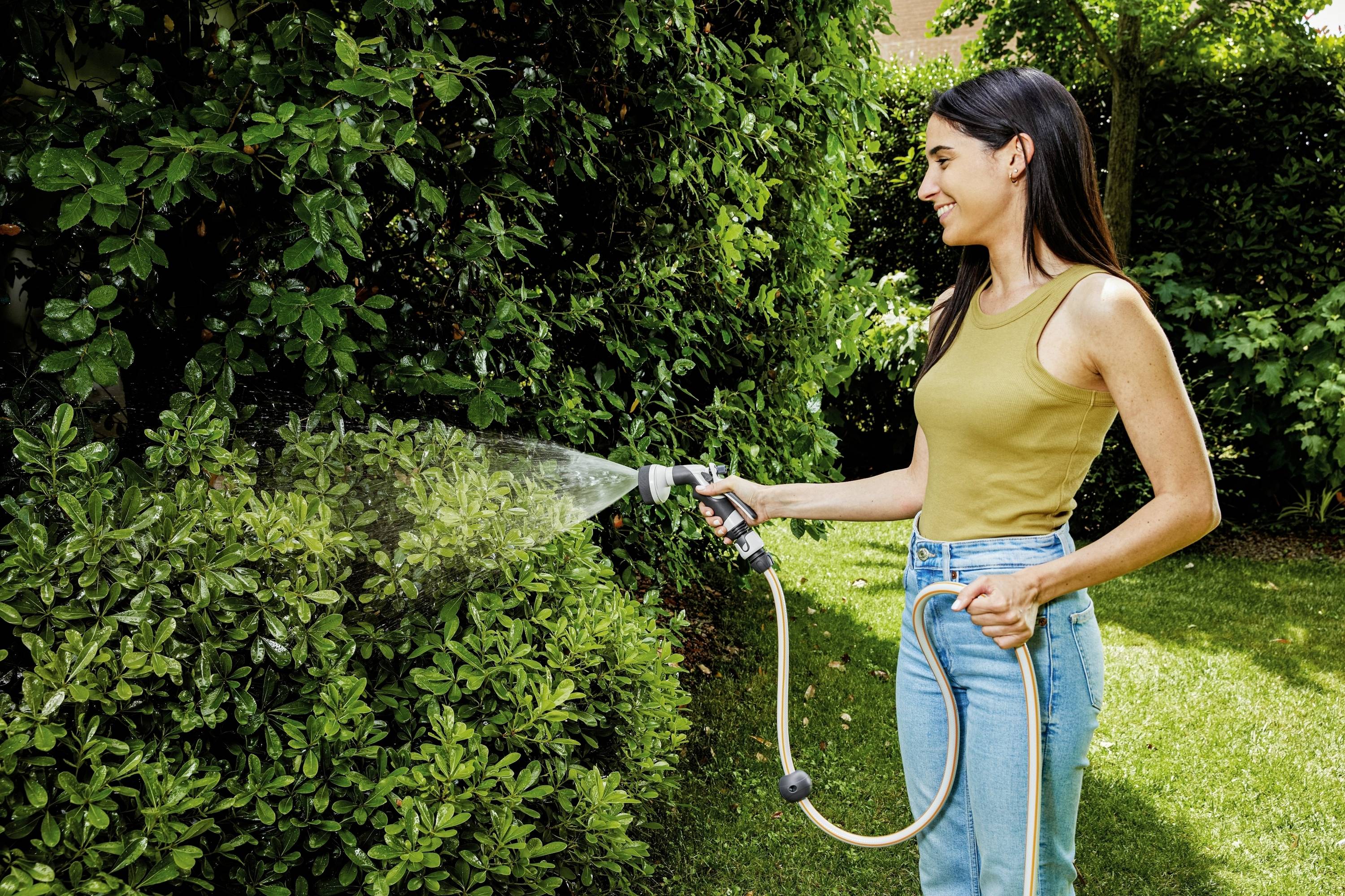A woman is standing in the garden, watering bushes with a hosepipe. She is wearing a yellow top and blue jeans. It is sunny.