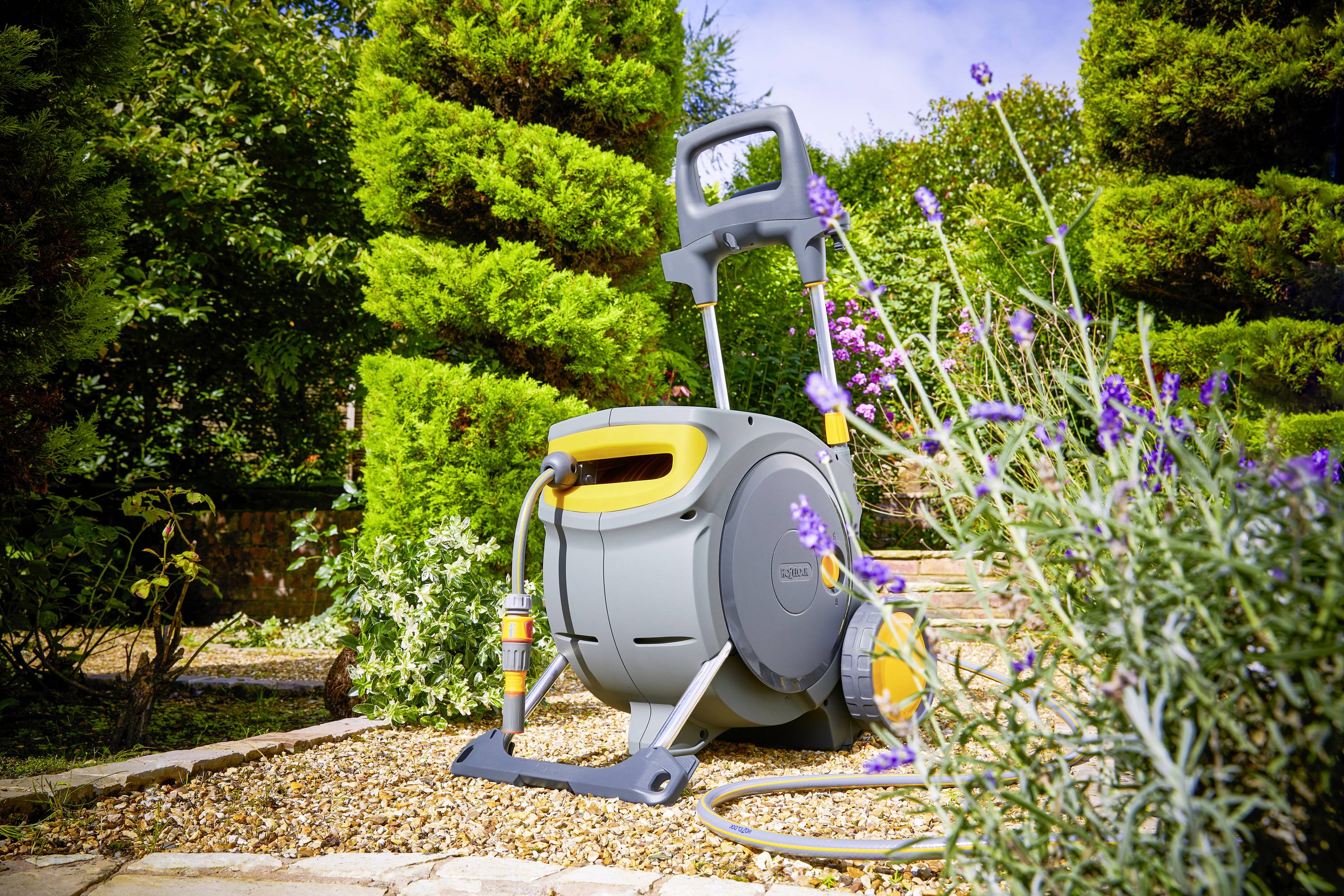 A garden watering can with a coiled hose stands in a well-maintained garden with flowers and trimmed bushes.