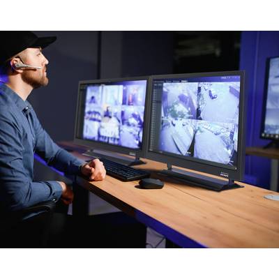A man wearing a headset sits in front of two monitors displaying surveillance camera images in a control room.