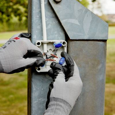 A person wearing grey gloves is installing an electrical cable in an outdoor electrical cabinet. Blue connectors are visible.