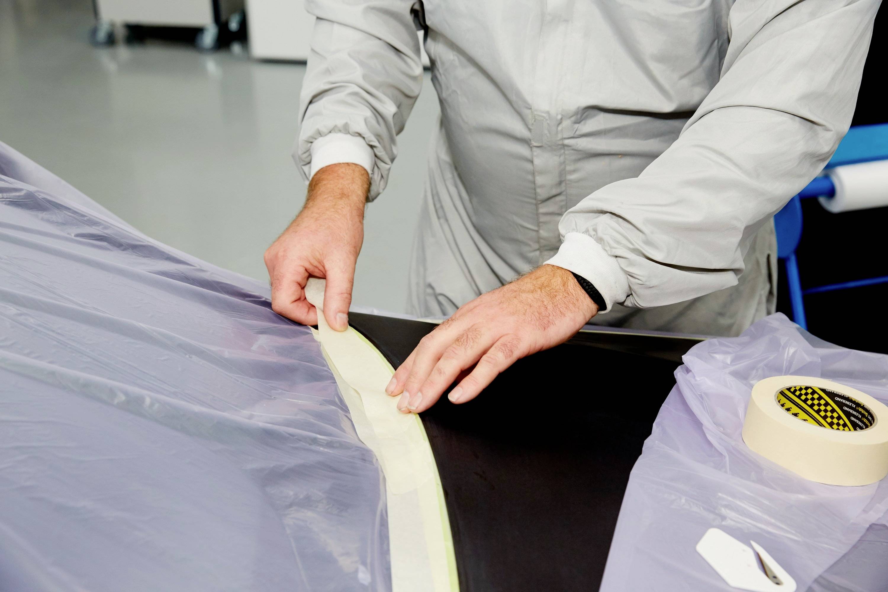 A person in protective clothing is applying masking tape to a plastic sheet. The scenario suggests preparations for a protective coating.
