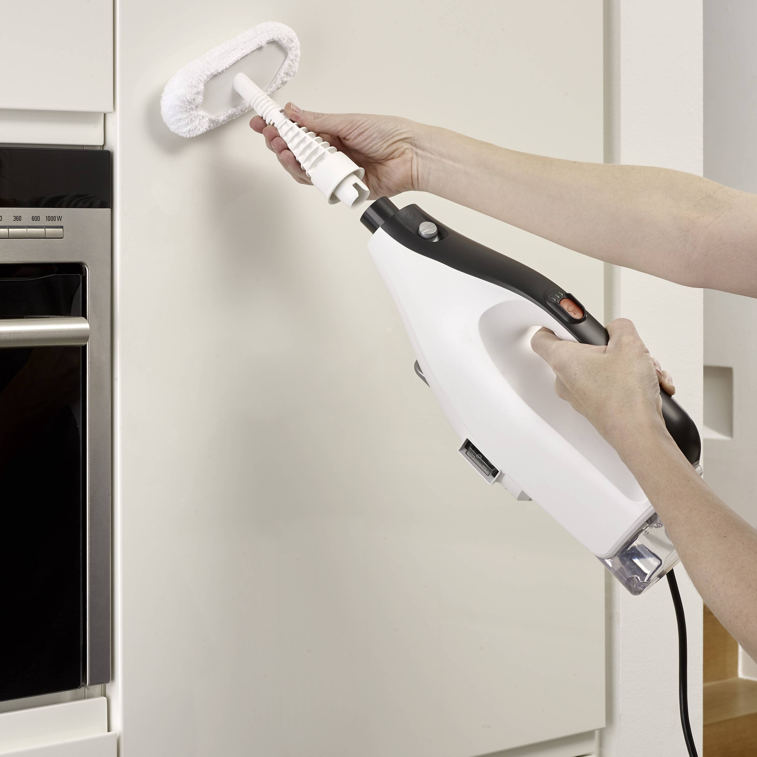 A person is cleaning a vertical surface with a steam cleaner. The focus is on the hand guiding the device.