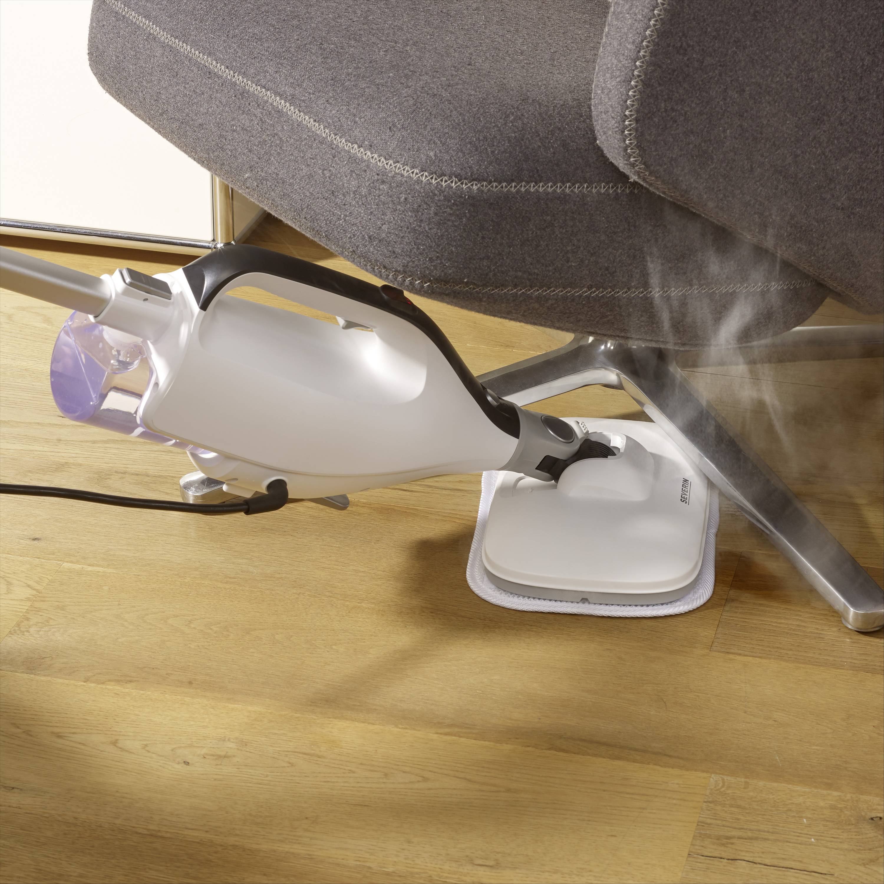 Steam cleaner generating vapour on a wooden floor beneath a grey chair. Carefully cleaning the floor with visible water vapour.