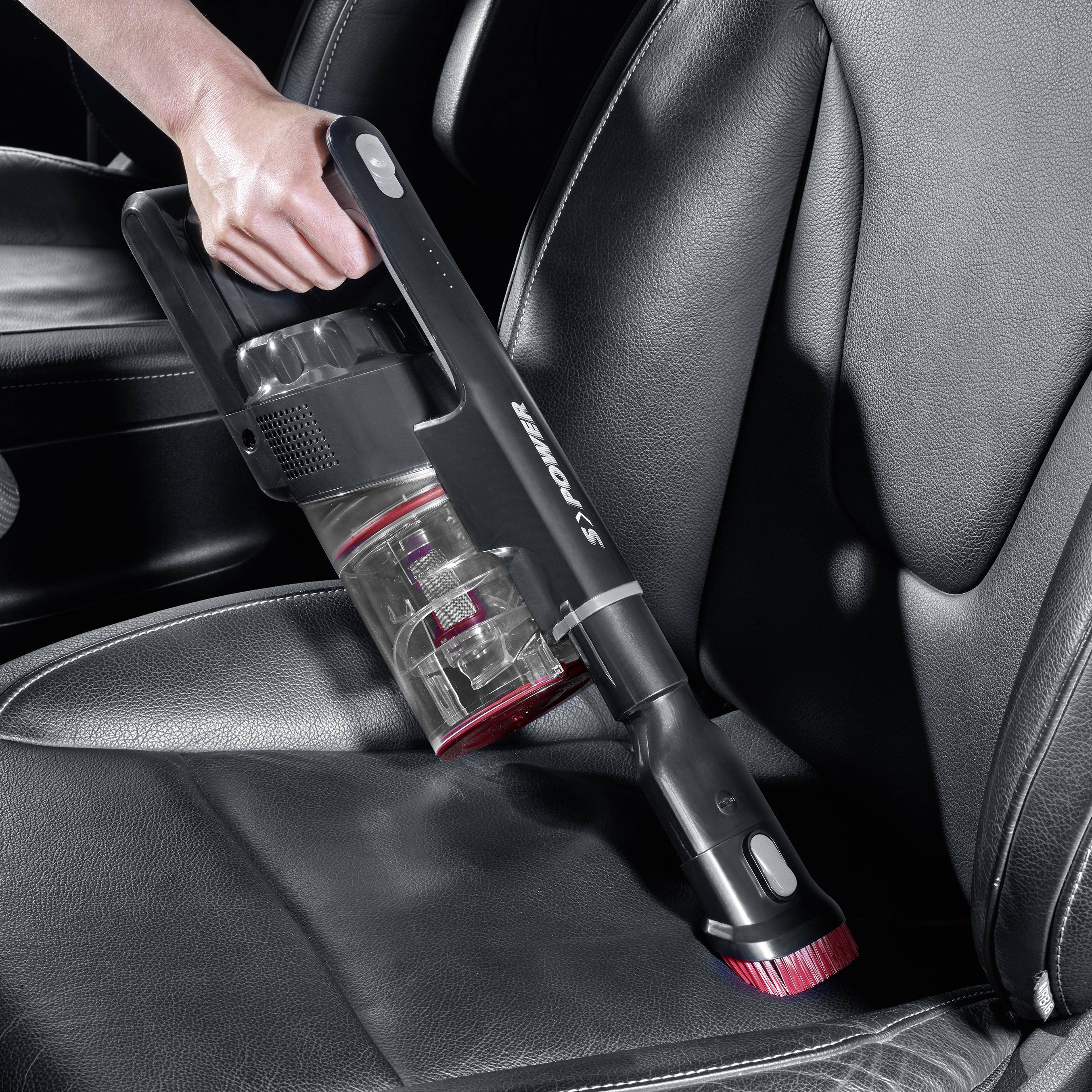 A person is using a handheld hoover to clean a car seat. The hoover has a transparent dust container.