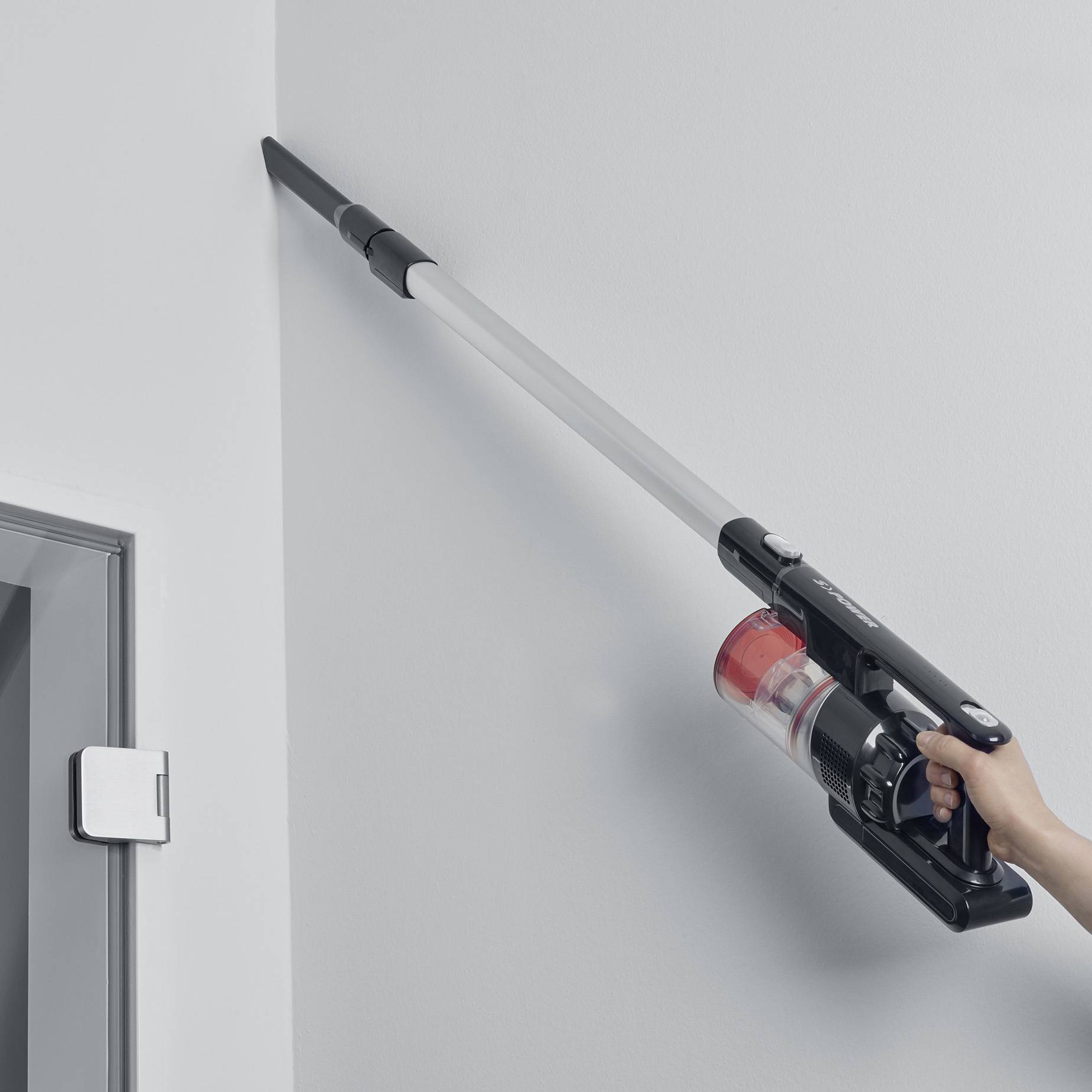 A person is holding a cordless vacuum cleaner and cleaning a high corner of a room. The vacuum cleaner is black and red.