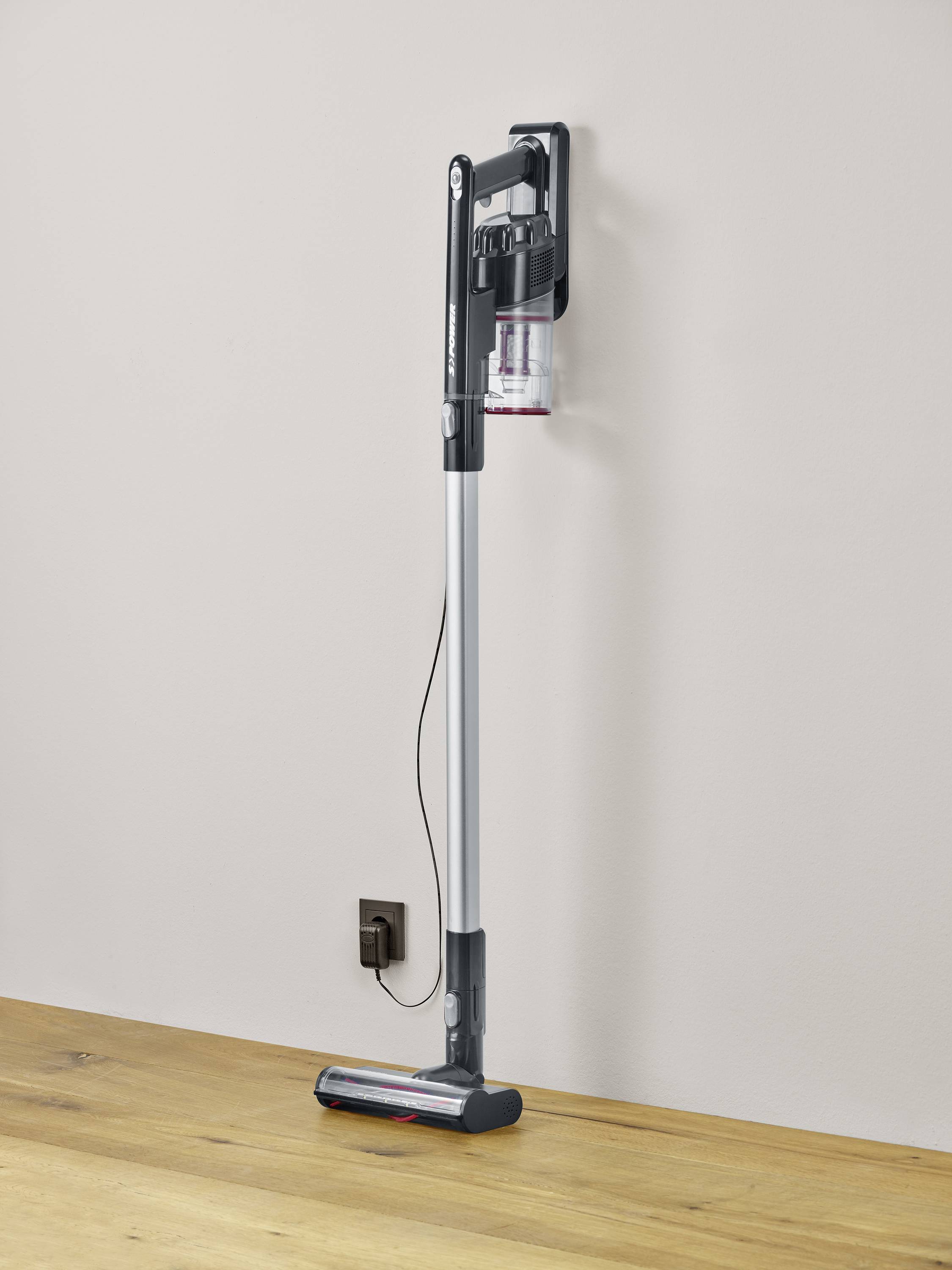 A slender cordless vacuum cleaner leaning against the wall on a wooden floor. The vacuum cleaner is black and silver.