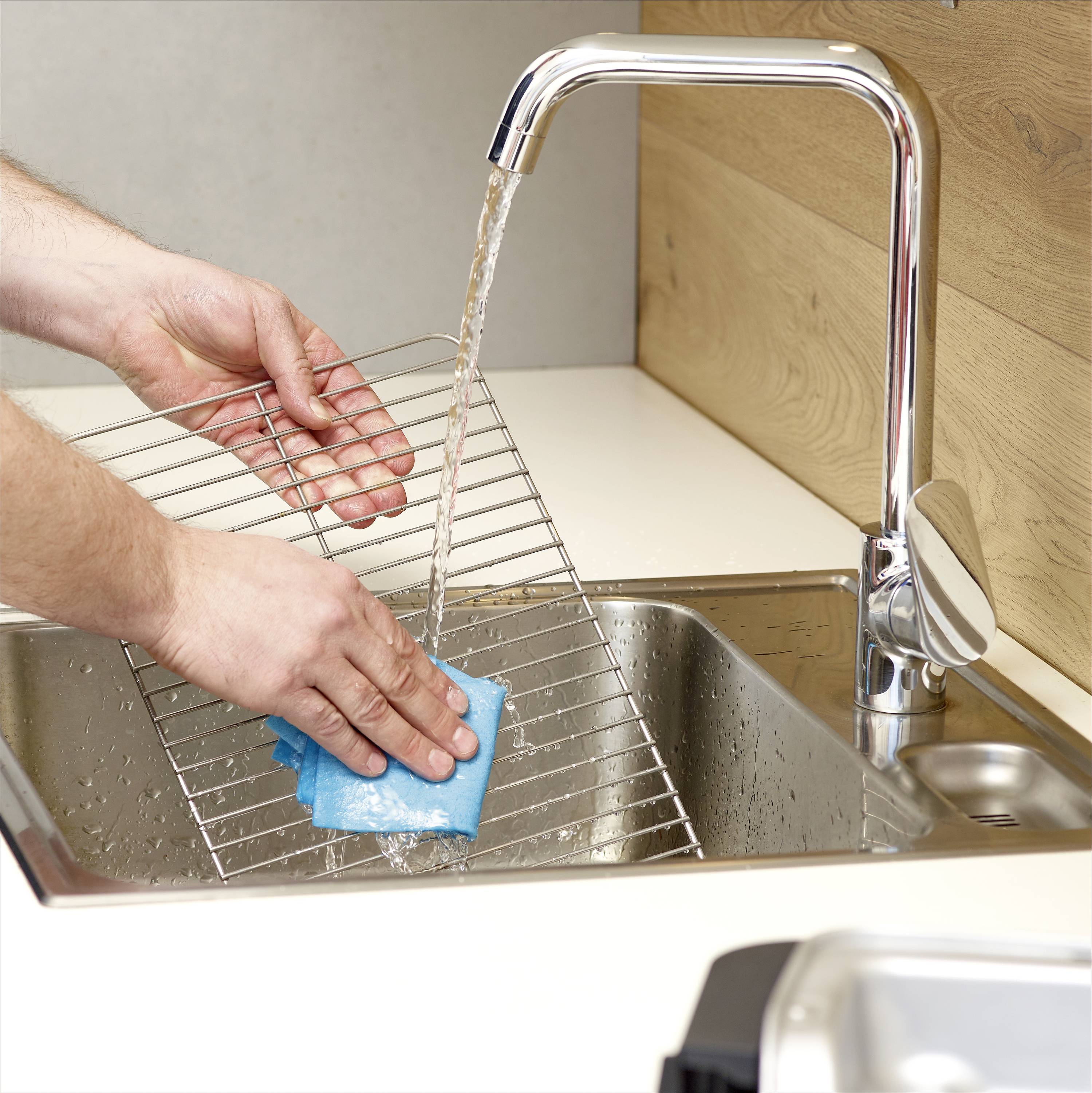 A person is cleaning an oven rack under running water with a blue sponge in a modern kitchen.