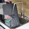 A person is cleaning a waffle maker with a green sponge under running water in a kitchen sink.