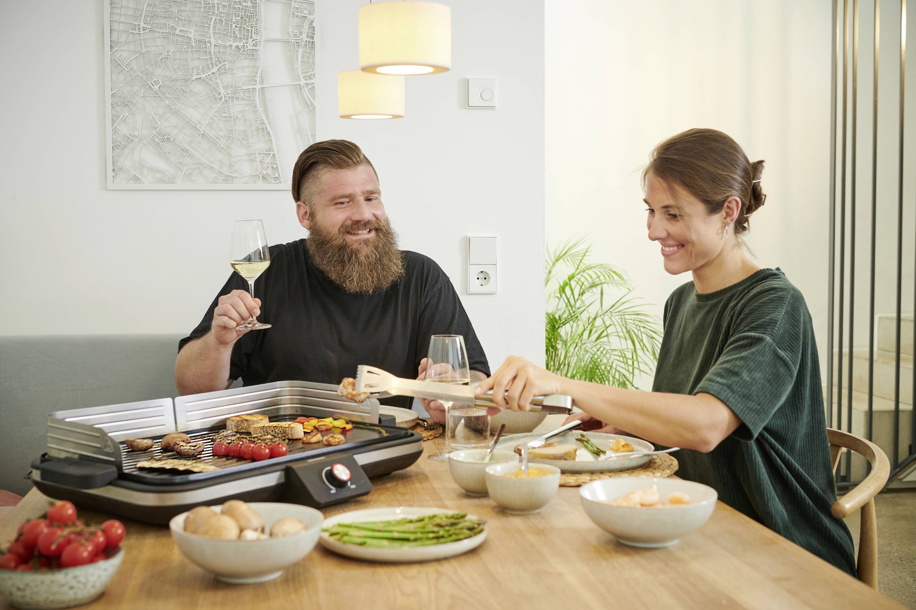 A man and a woman are sitting at a table with an electric tabletop grill. They are laughing and raising their glasses.