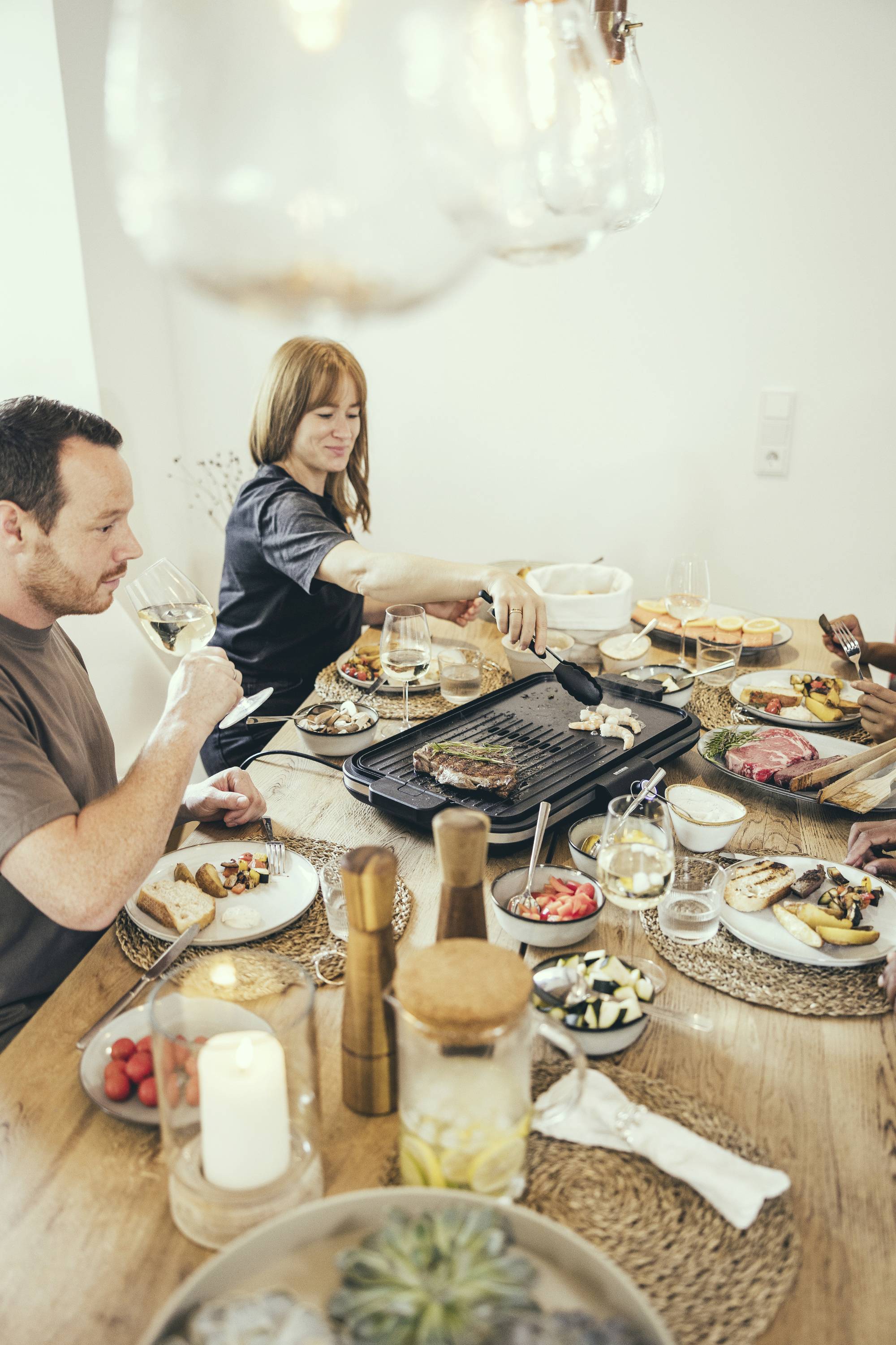 A man and a woman are sitting at a set table eating. The woman is operating a grill plate, while the man holds a glass of wine.